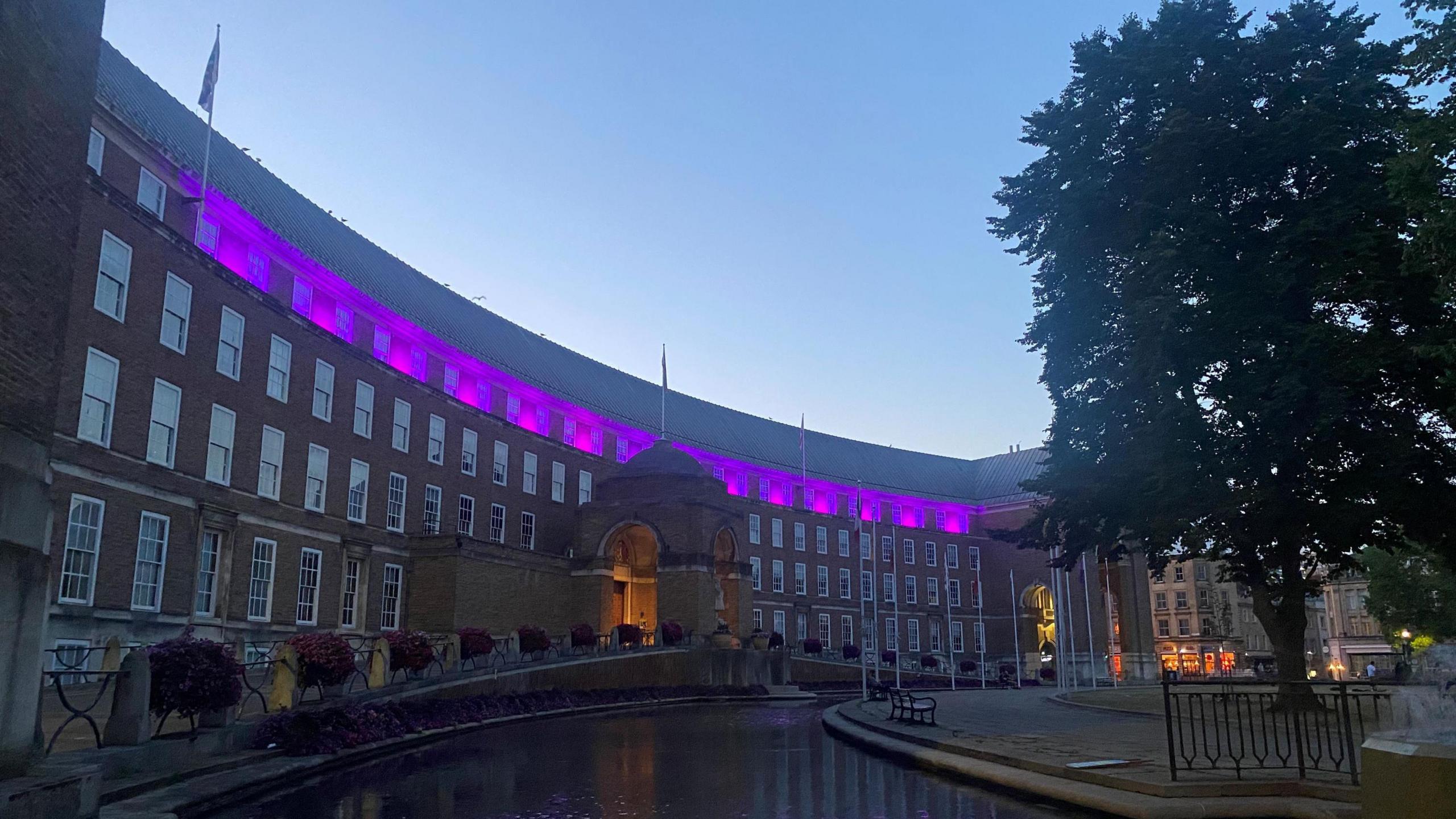 City Hall on College Green in Bristol is seen illuminated by purple lights along its top floor. The picture is taken after dark