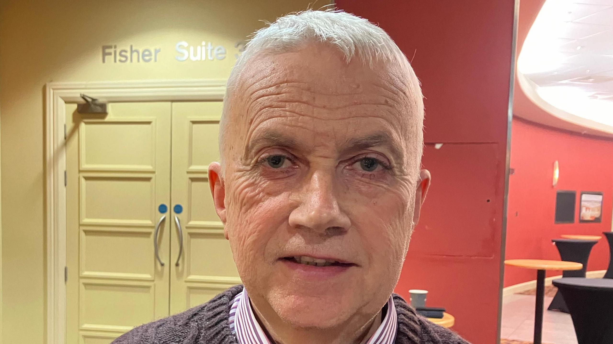 Francis Gallagher is wearing a striped shirt with a patterned jumper. He has grey hair. Behind hi to the left is a cream wall with double doors, the words Fisher Suite is above the doors in silver font. To his right is a red wall with tables along it. 