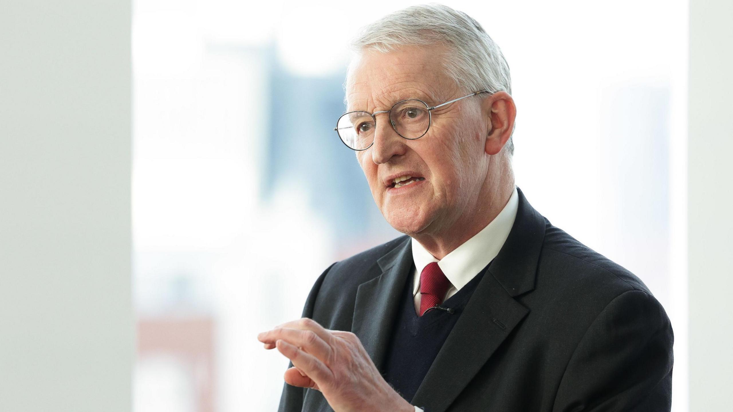 Hilary Benn wears a black suit, black waistcoat, white shirt and red tie. He has grey hair and wears glasses.