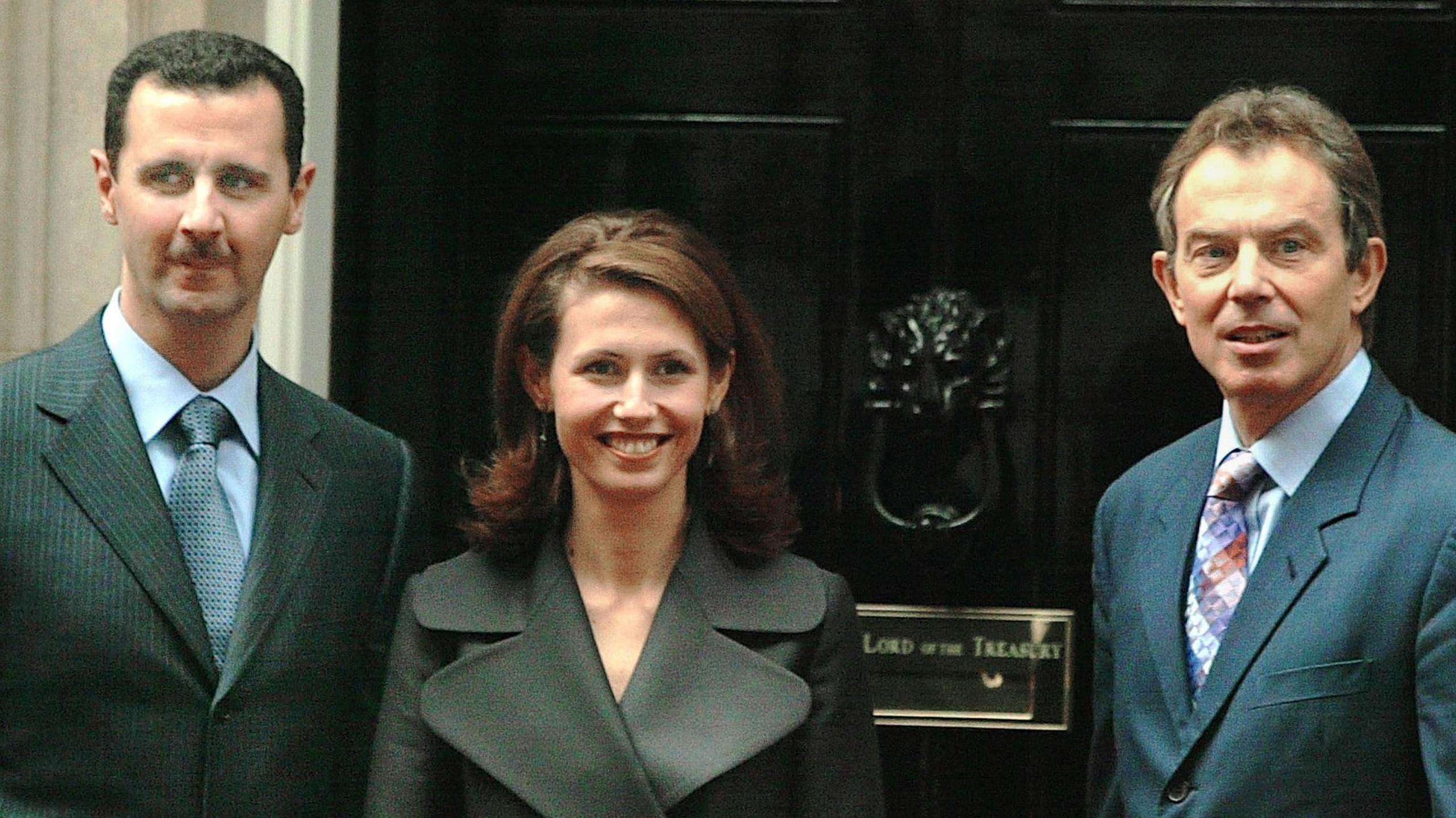 Bashar al-Assad on the left wearing a grey suit, stands beside Asma al-Assad, who is smiling widely in a grey coat. Tony Blair stands beside them both, with the black door of Number 10 Downing St behind them.