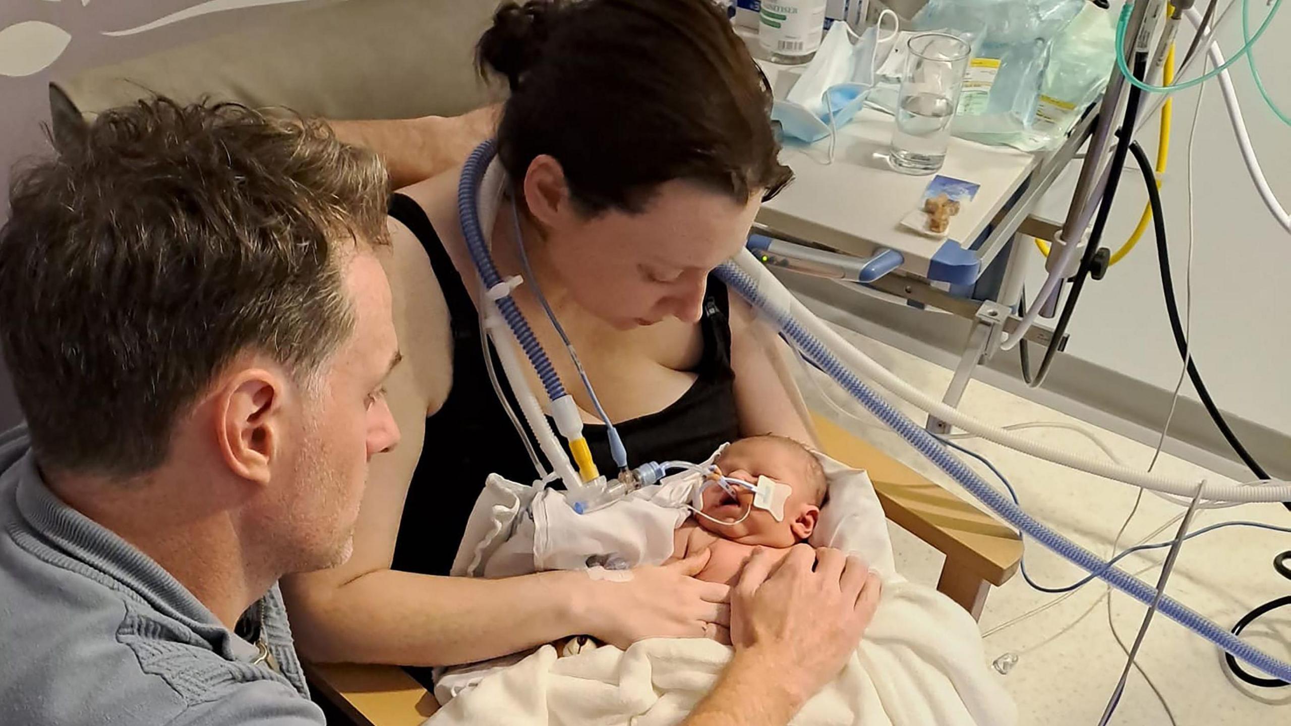 Rhodri Thomas ac Emily Brazier mewn ward mamolaeth, yn edrych ar eu babi newydd-anedig. Mae Liliwen ym mreichiau ei mam ac mae sawl tiwb yn ei thrwyn.