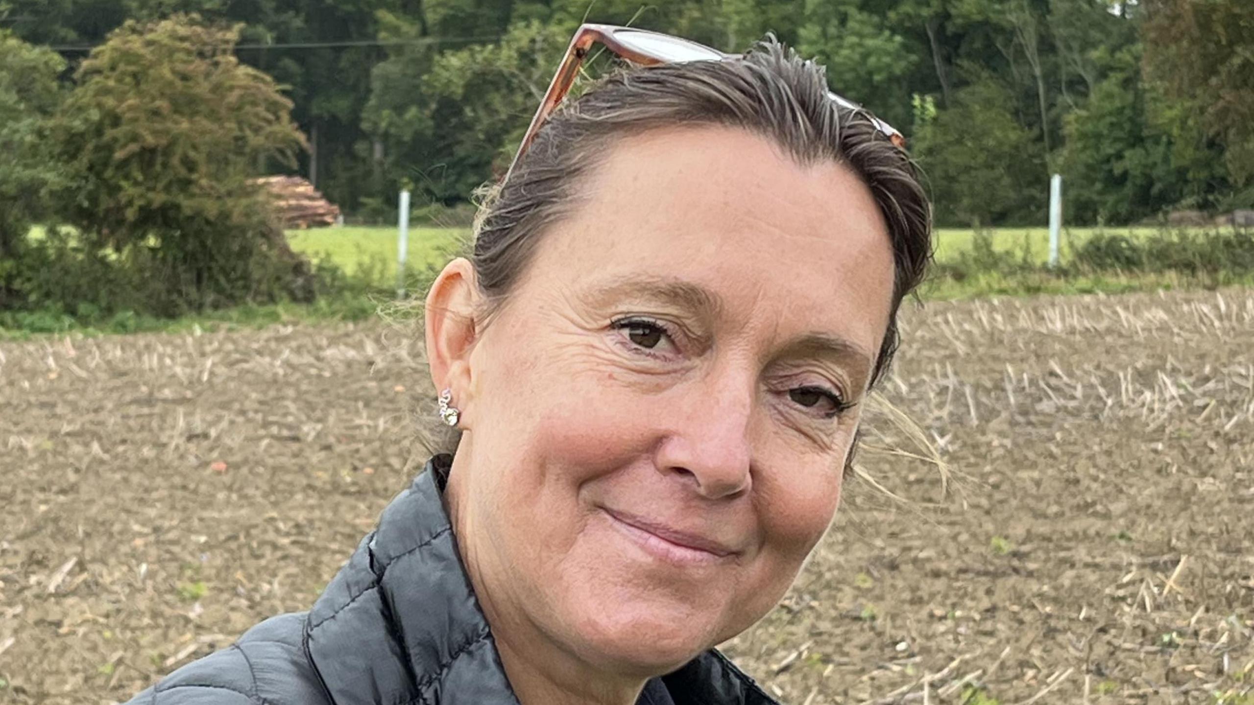 A woman with brown hair smiling at the camera. She is standing on a brown field.