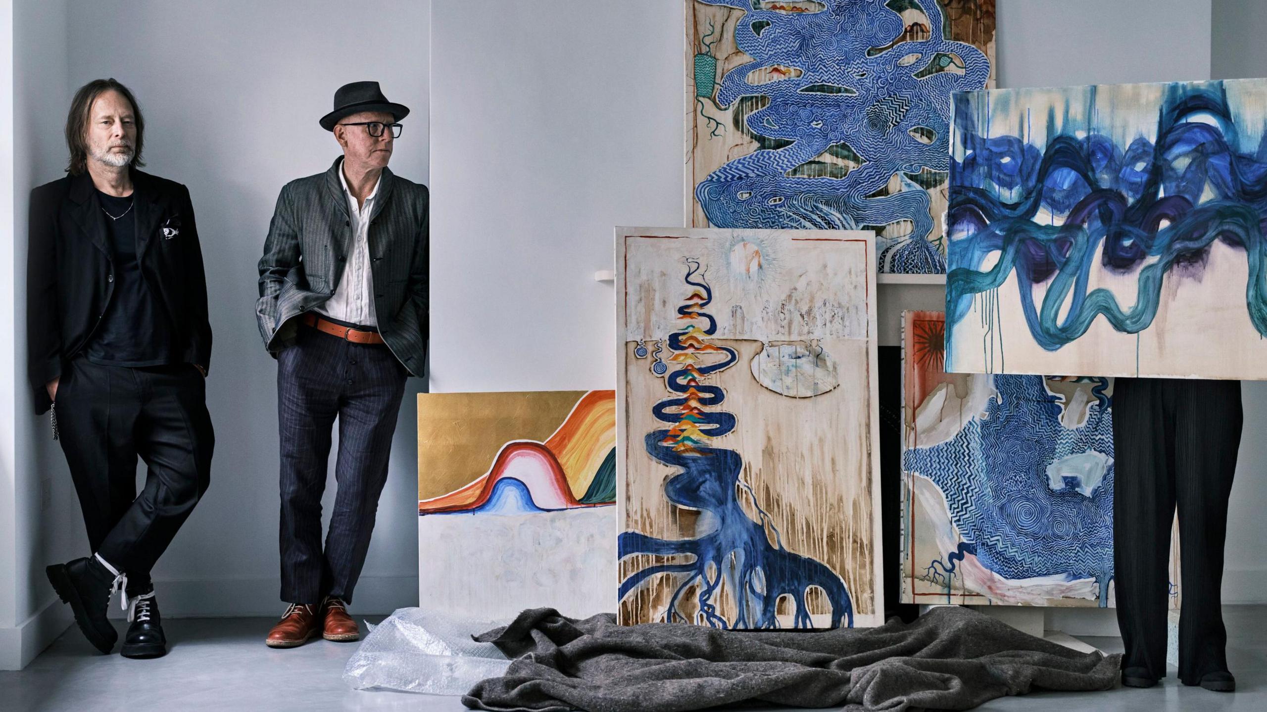 Thom Yorke and Stanley Donwood lean against a wall in a white room next to some of their artwork. Someone's legs can be seen below one painting as the person holds it aloft.