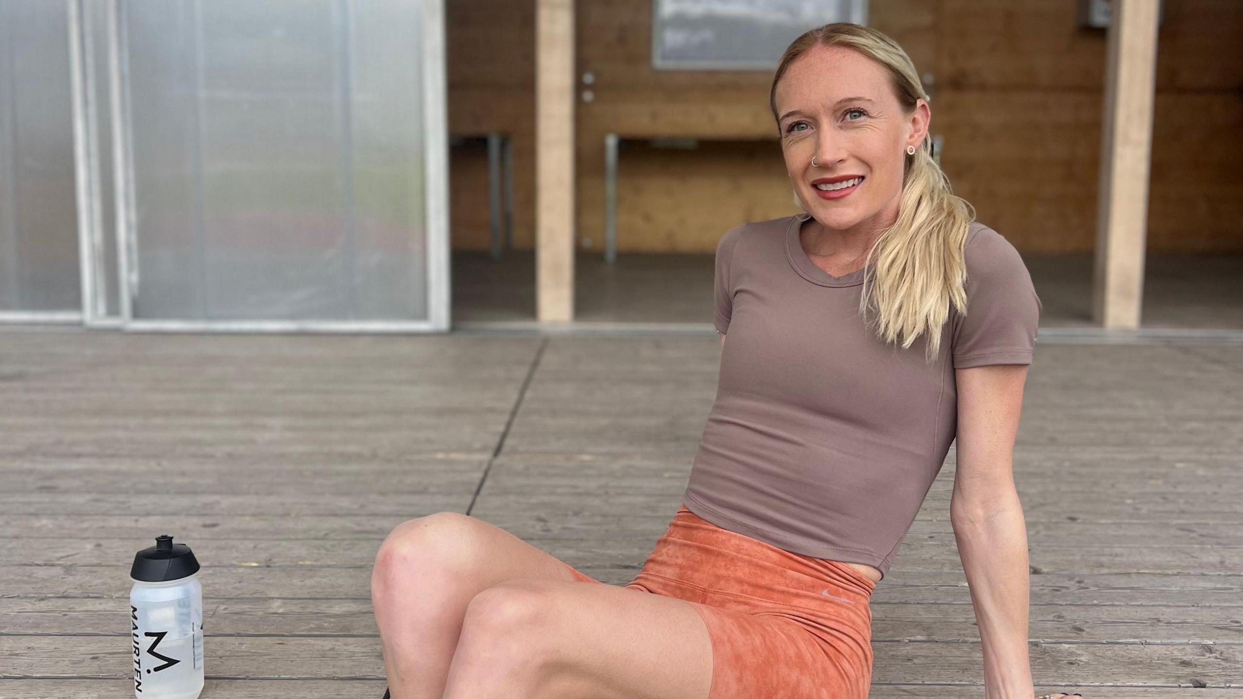 Calli with the slender frame of a distance runner, blonde hair over one shoulder, blue eyes, red lipstick and sat on wooden decking leaning back on her hands.  