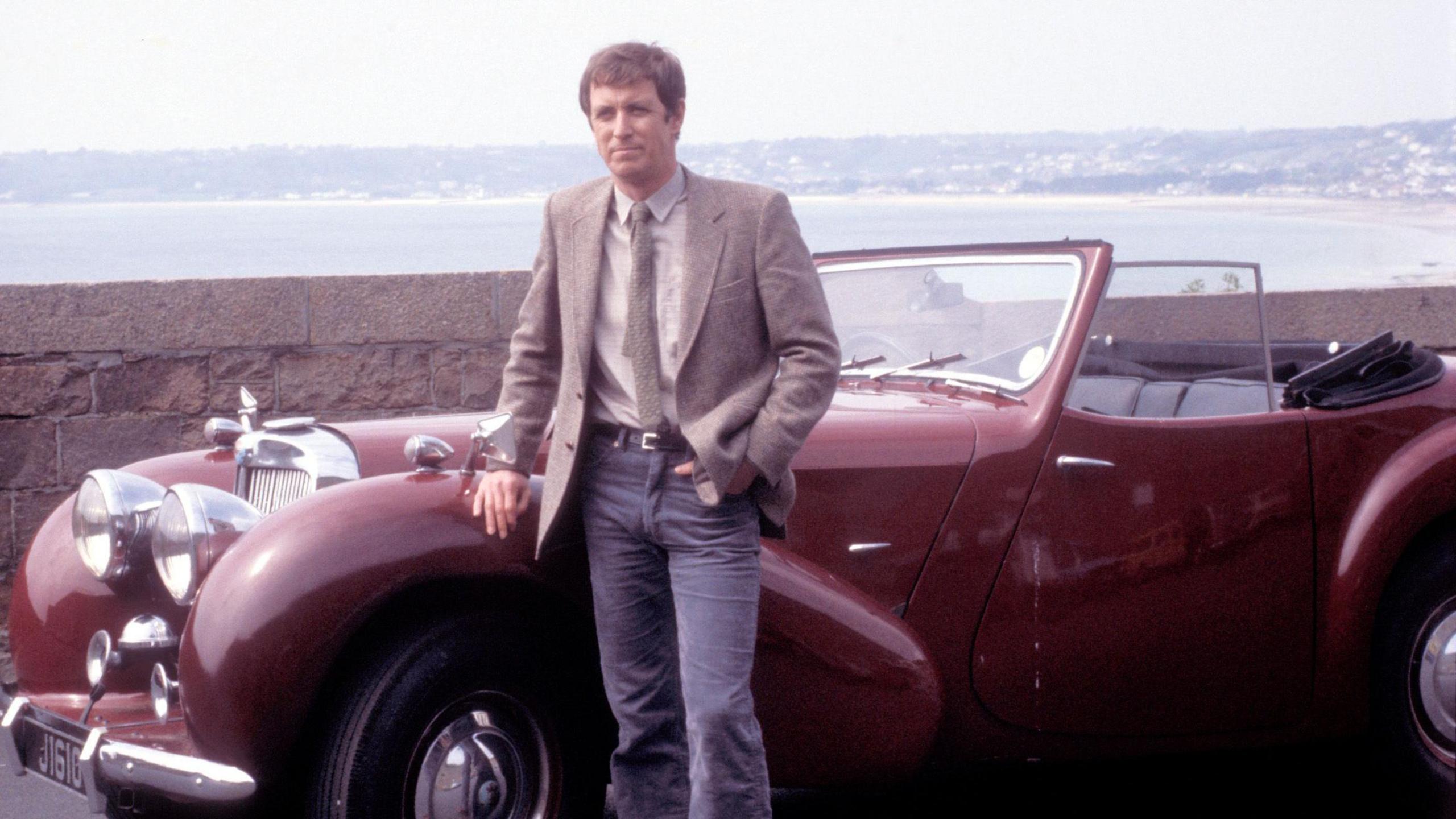 John Nettles pictured as Bergerac in the original series. He is seen learning against a dark red classic car while wearing a green tweed jacket with a beige shirt and green tie.