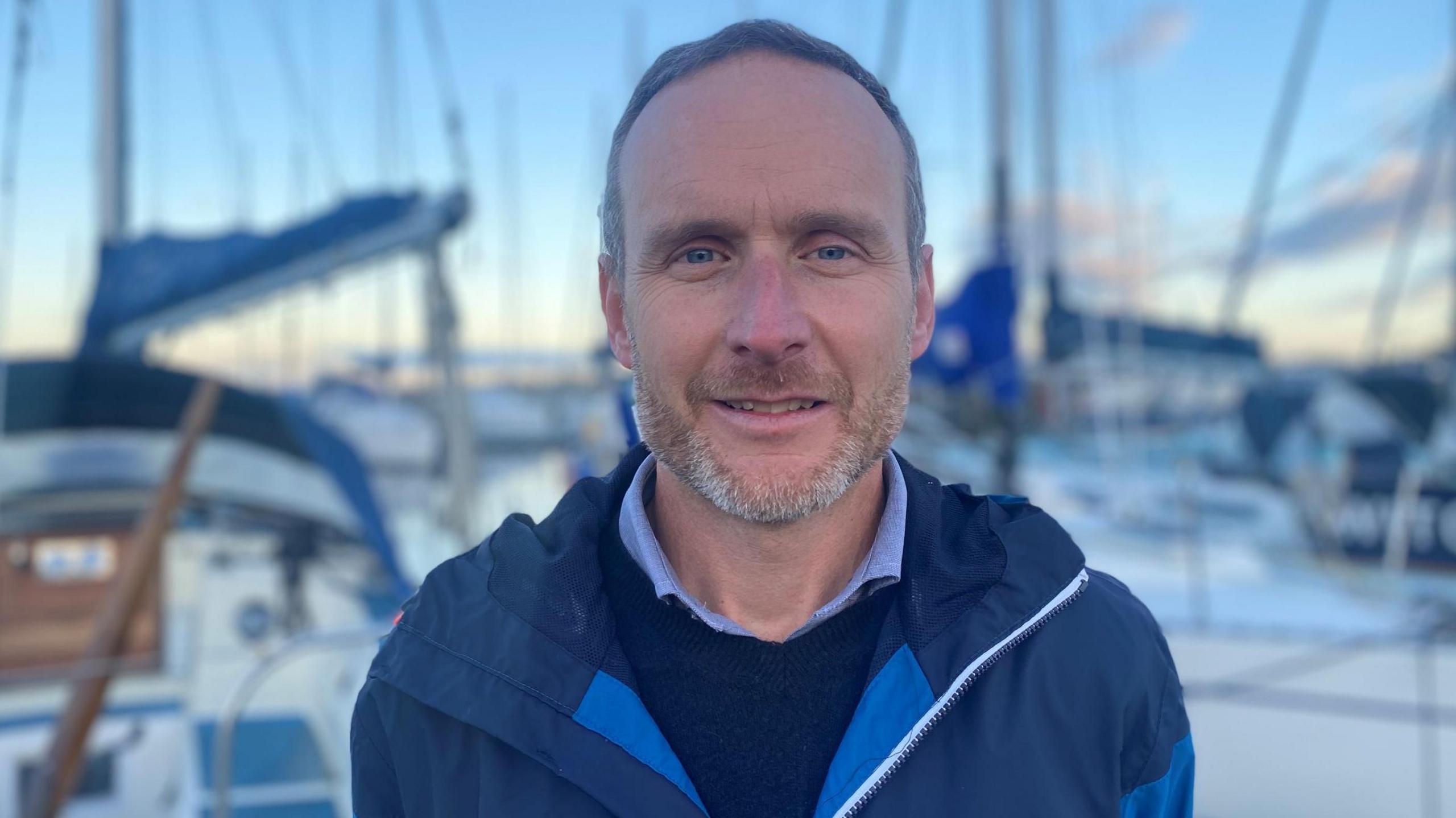 Stuart Fox is at Nairn harbour and there are boats and masts behind him. Stuart has short fair hair and designer stubble. He is wearing a blue jacket over a blue jumper and shirt.