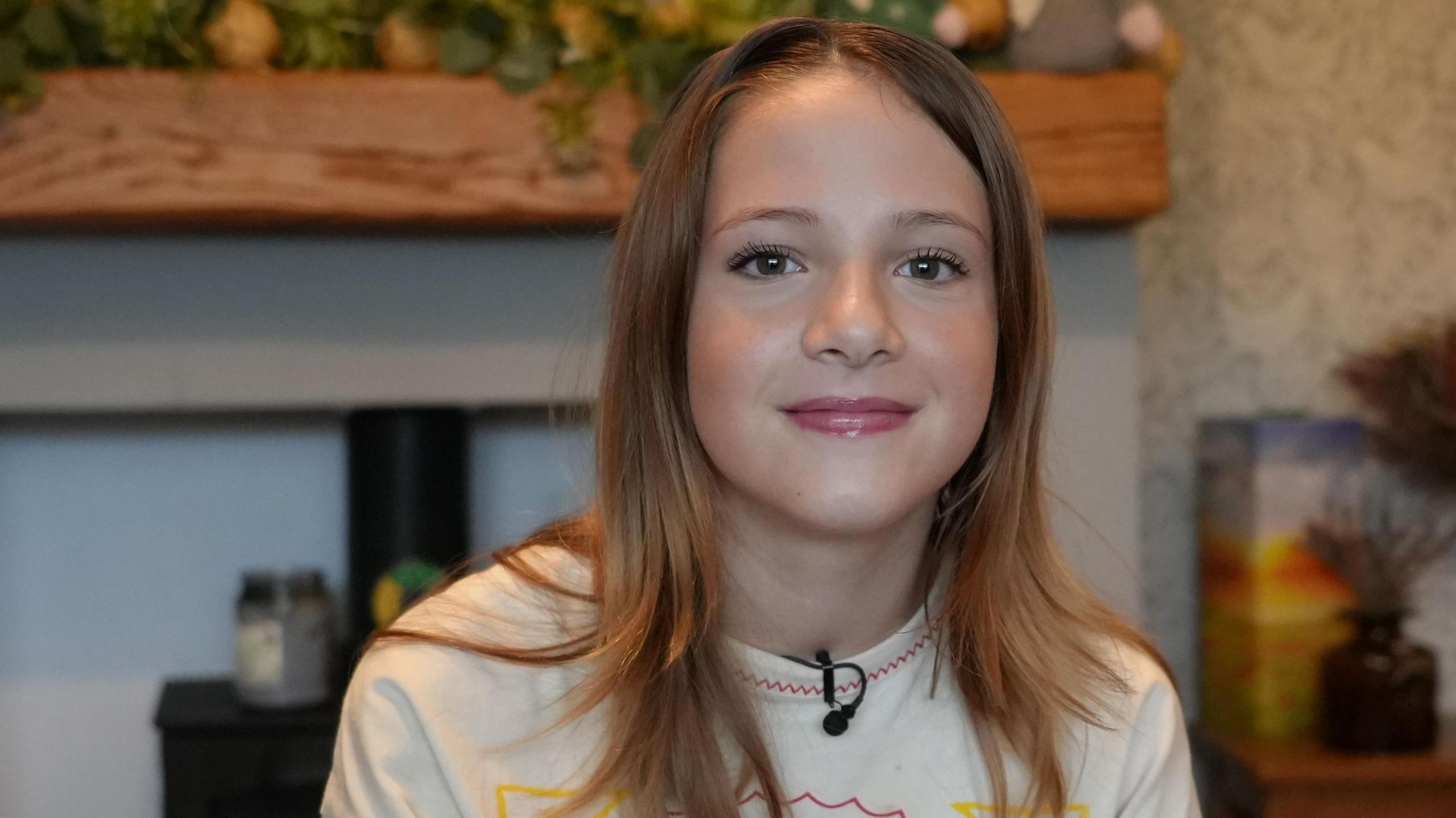 A 13-year-old girl with long straight mousy hair, and wearing a white patterned t-shirt, smiling to camera. 