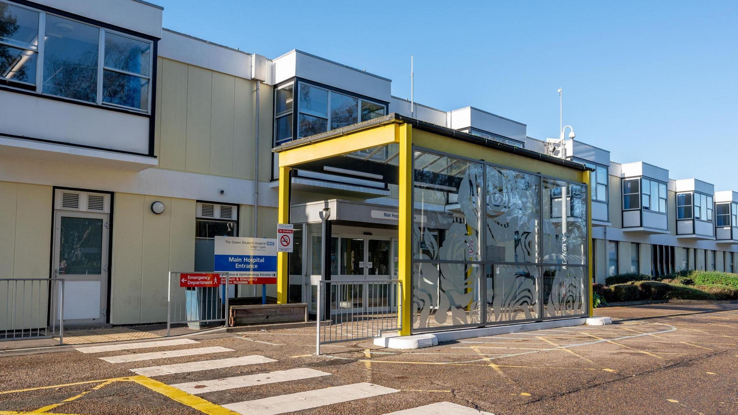 Exterior of Queen Elizabeth Hospital showing a two storey, low-lying building with first floor windows jutting out and a yellow framed and covered entry to the double doors