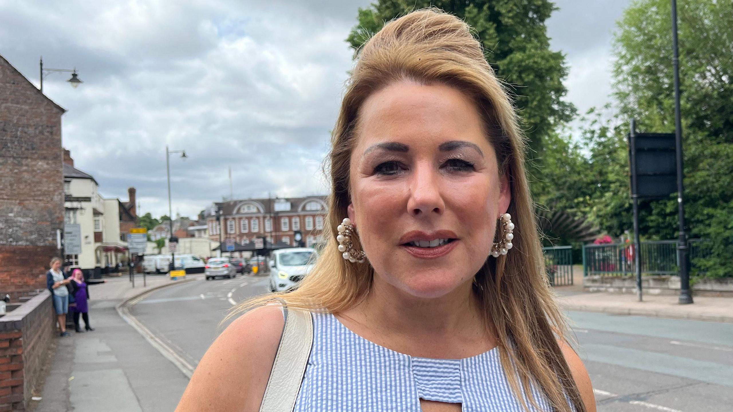 Siobhan smiling slightly and looking into the camera. She's wearing a light blue and white striped dress and is stood on a pavement with the road visible behind her with some trees.