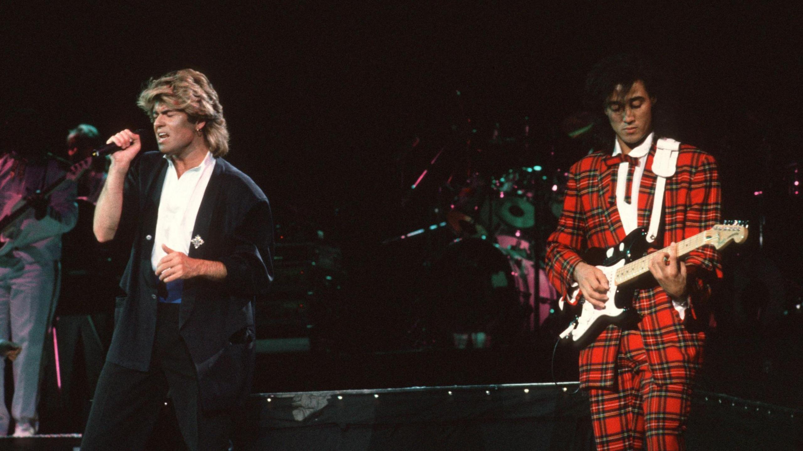 Wham! perform on stage during a concert. Michael, who has blonde highlights and is wearing a black suit and white shirt, sings into a microphone. Ridgeley, who has a black floppy fringe and is wearing a red tartan suit and white shirt, plays an electric guitar.