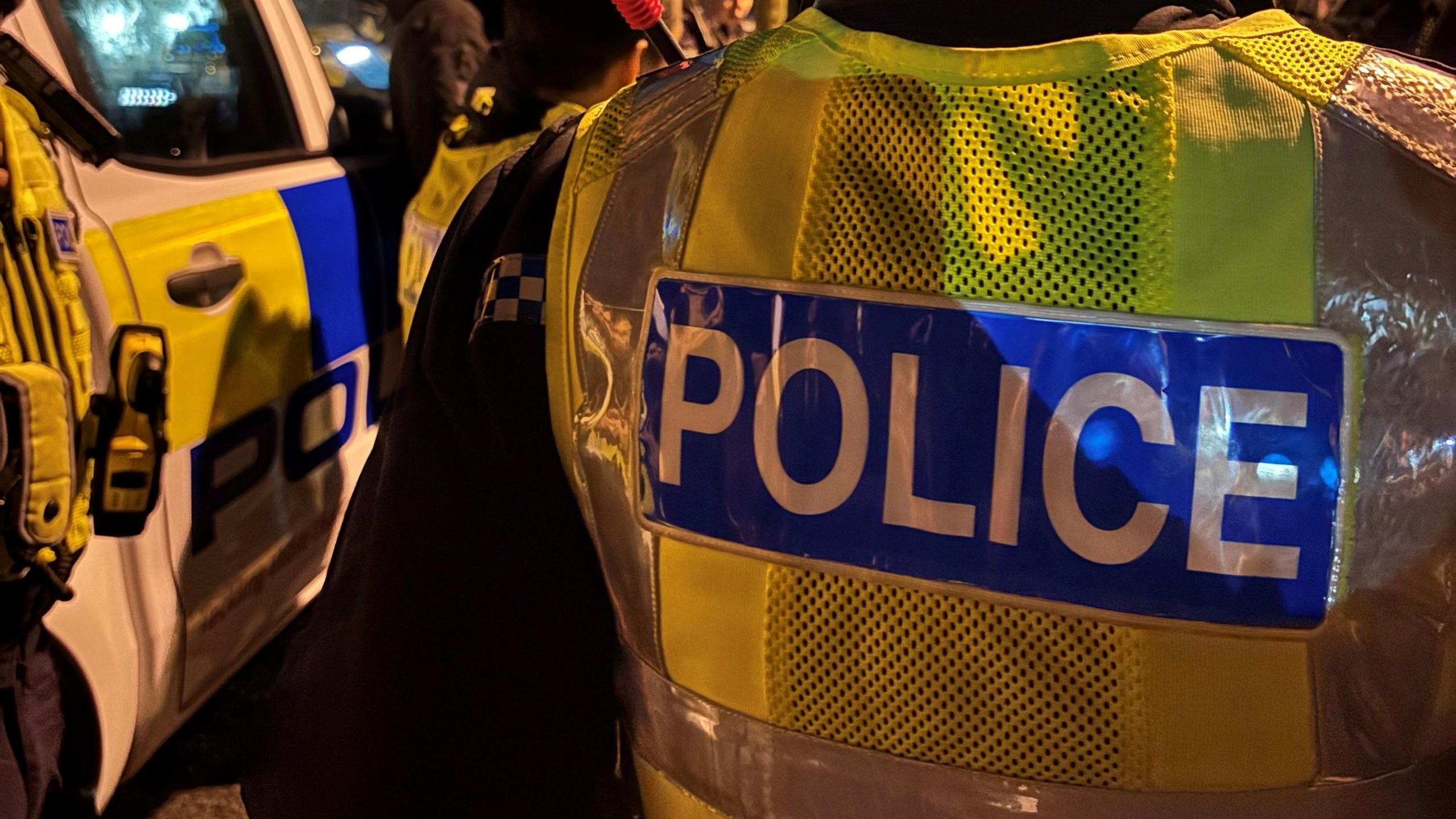 The back of a police officer who is wearing a yellow fluorescent jacket which says POLICE in white writing in a blue box. The picture has been taken while it is dark outside and a police car can also be seen. 