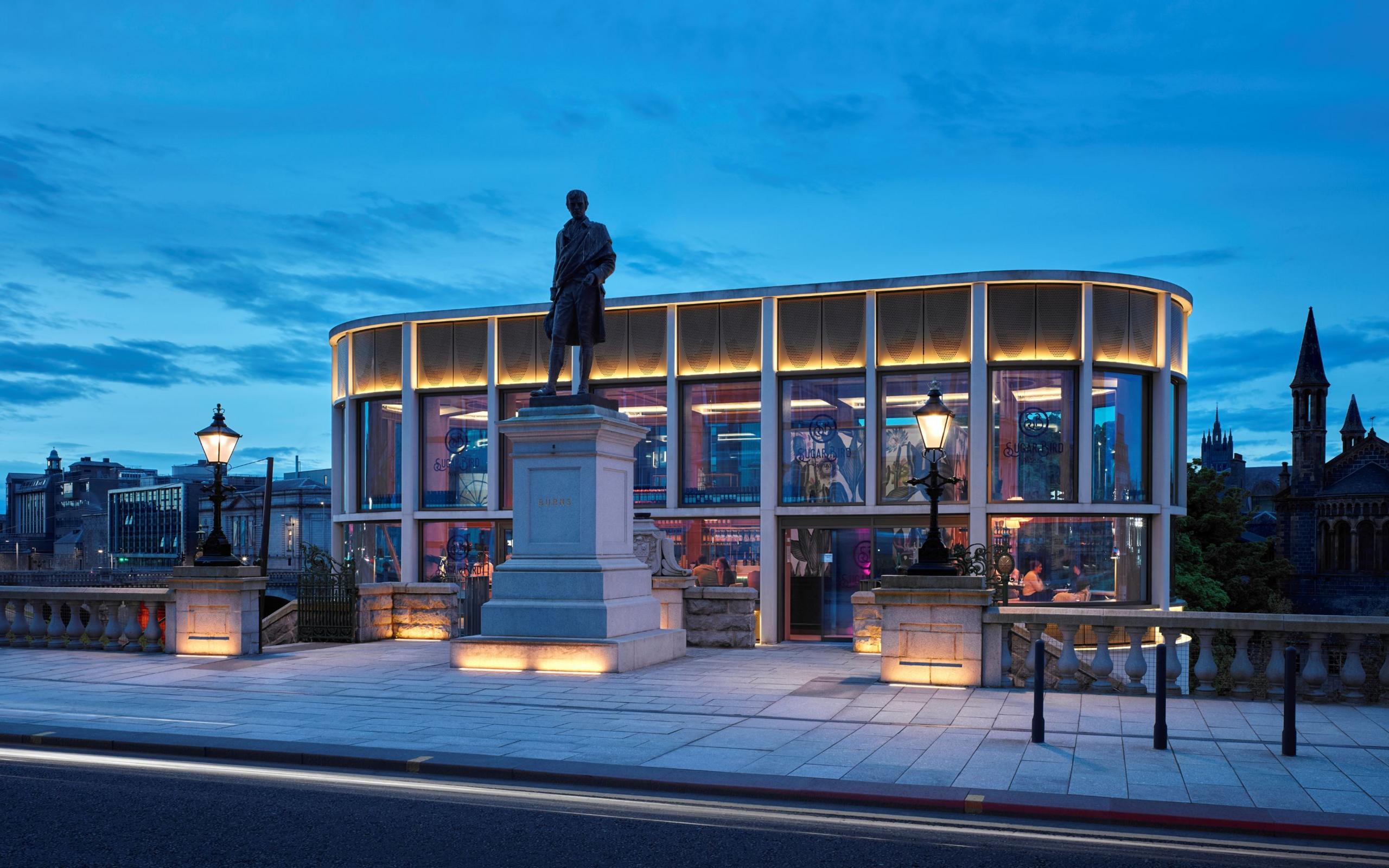 A view from the road of one of the pavilions - a long rounded-end glass box of a building, with warm lighting, containing a wine bar. A lit-up statue and street lamps sit outside and stone balustrade runs along either side of the building.