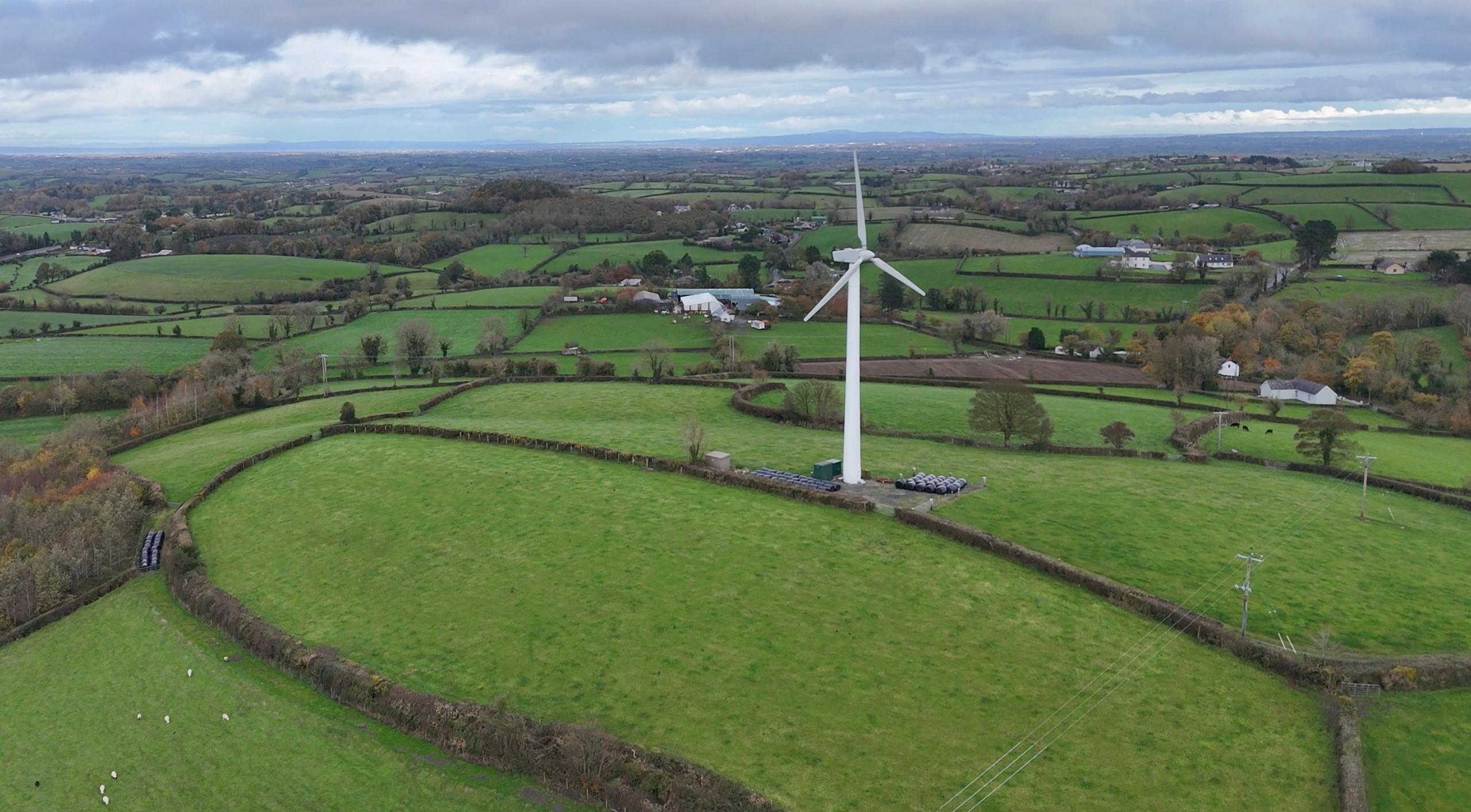 An aerial picture of white turbine stands on top of a hill. It is surrounded by green fields separated by low hedges, with grazing sheep visible in one of the fields.