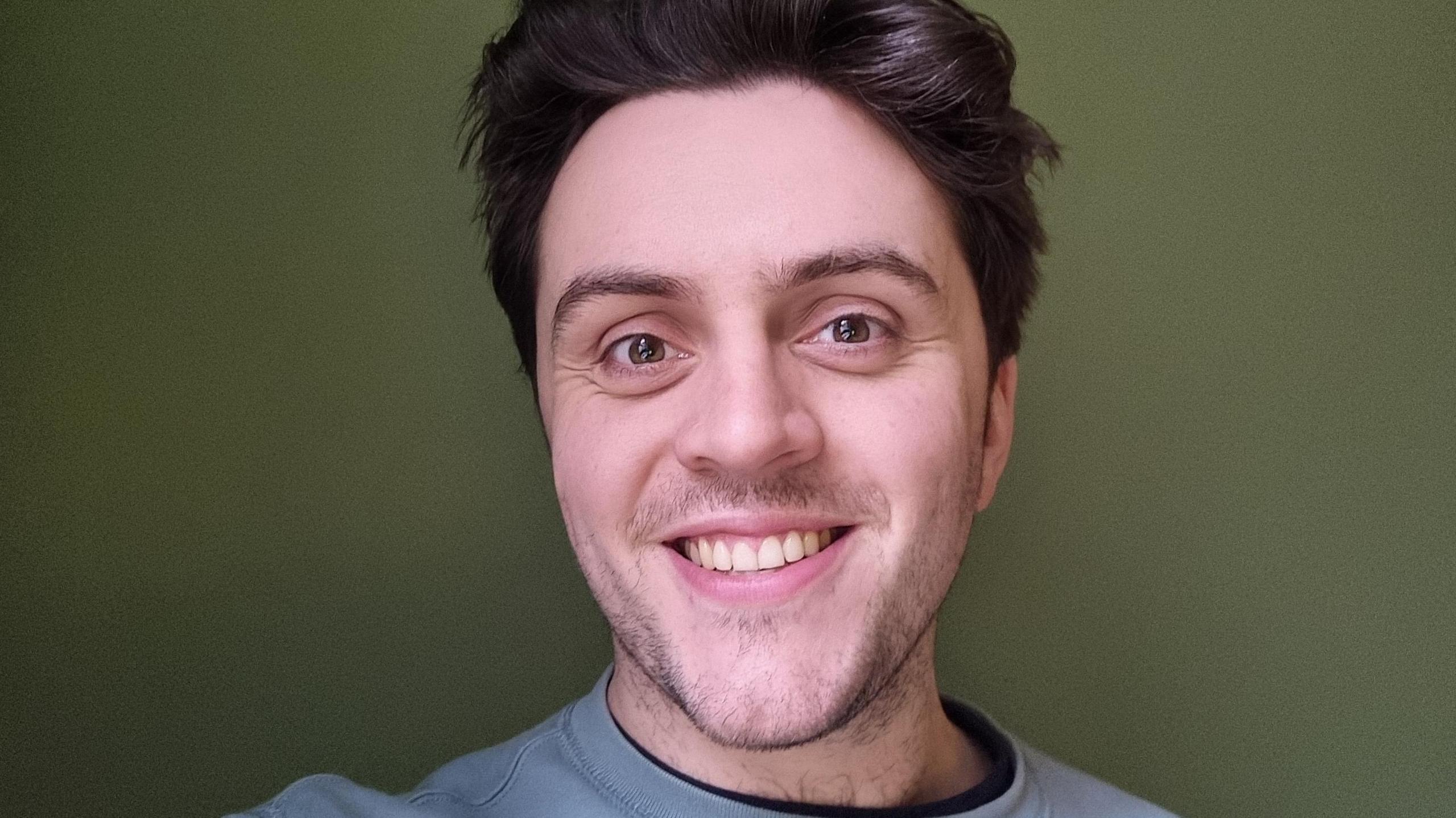 A man with brown hair and brown eyes smiles at the camera. He is wearing a blue jumper and standing in front of a green wall.