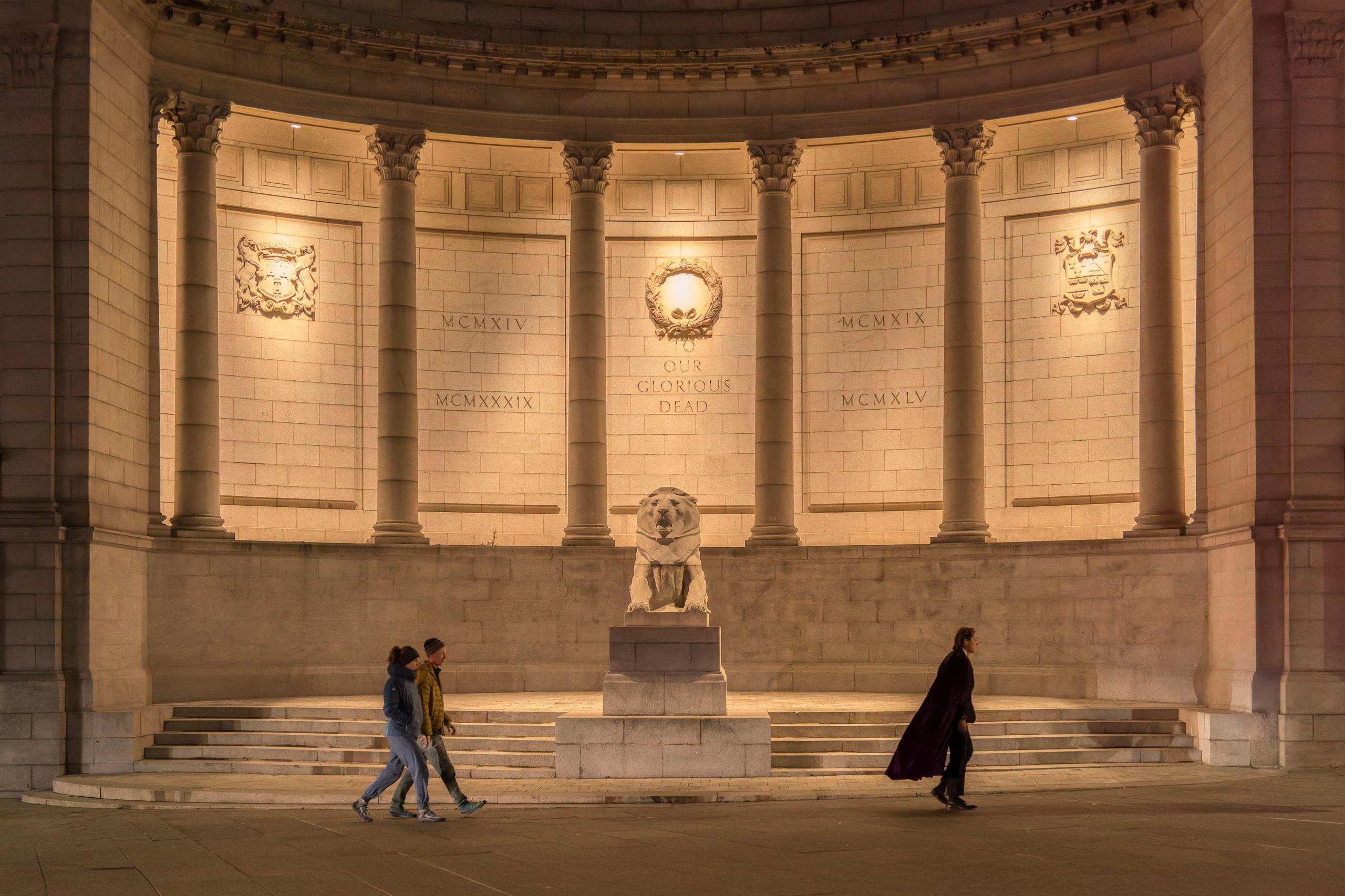 There is a lion statue. Behind it are pillars and a white wall that is lit up.