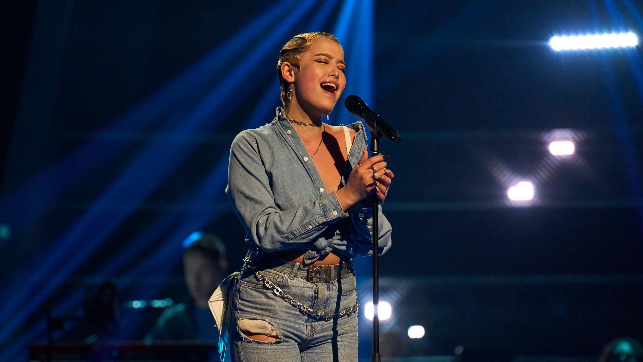 Ace, wearing jeans and a denim shirt, singing on The Voice. She has short blonde hair, and out of focus behind her is a man playing the piano