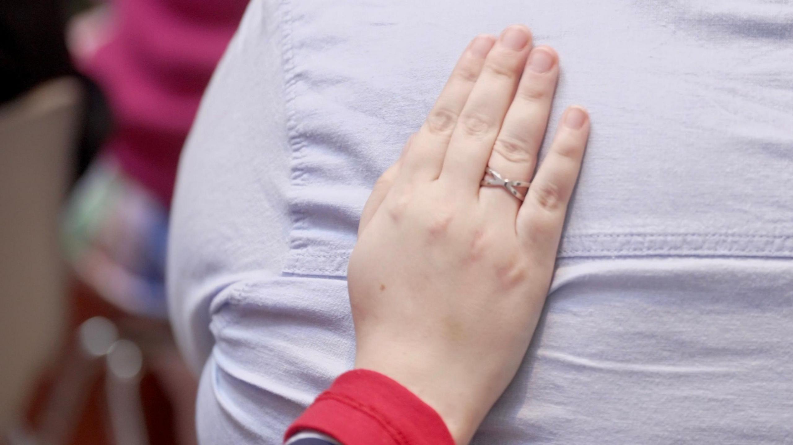 A woman's hand with a ring on it reaching out at a support group meeting to rest on the shoulder of the person in front, in an apparent gesture of reassurance.
