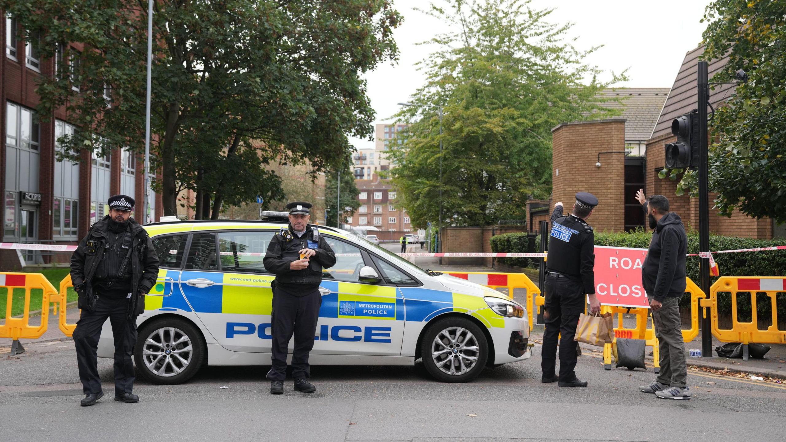 Three police officers stand in front of a police car and police cordon at the scene of a fatal shooting. One officer speaks to a member of the public.