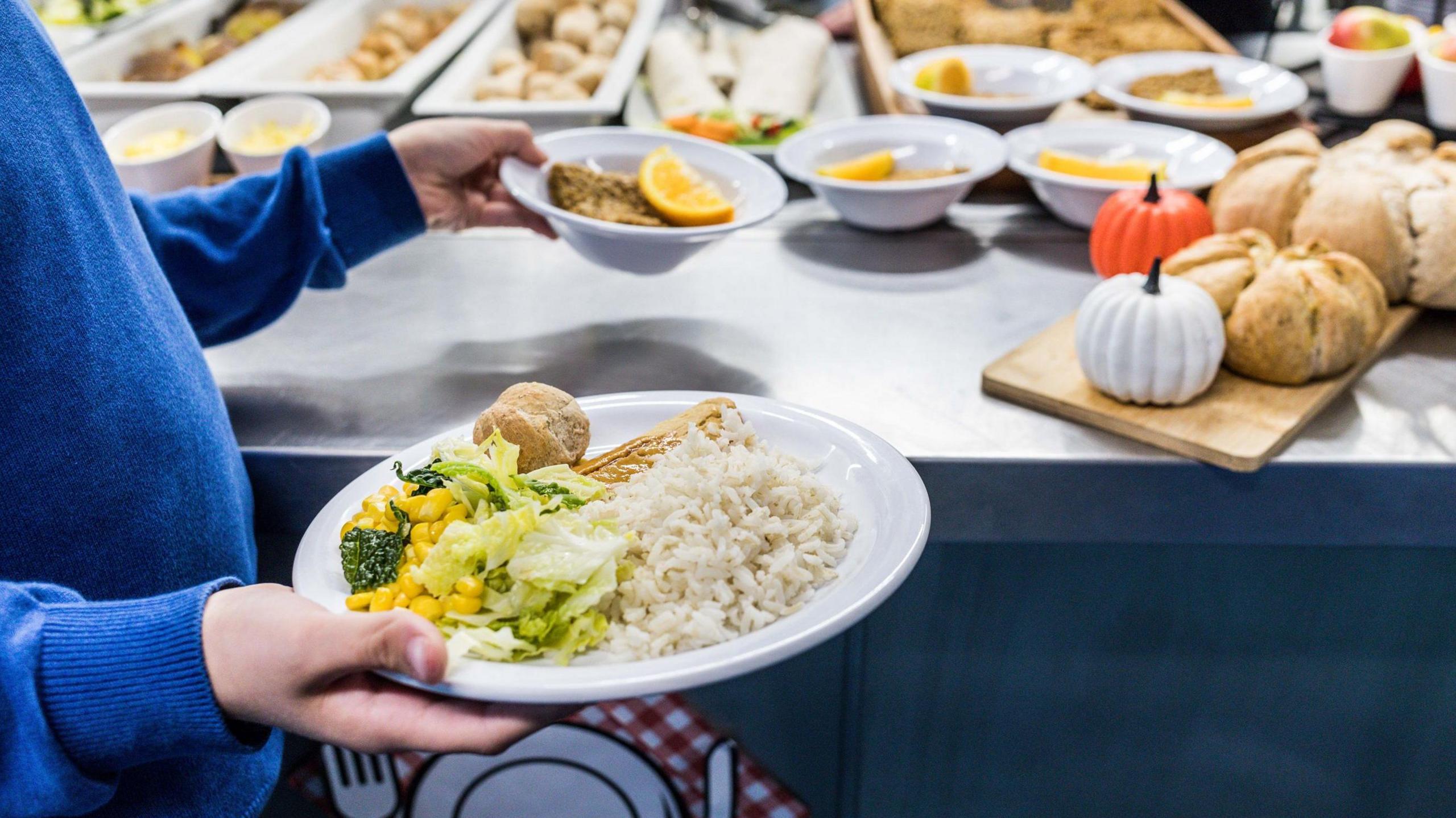 A woman holds a plate with rice, salad and bread and a bowl with orange and cake. There is a table behind her with more plates full of food.