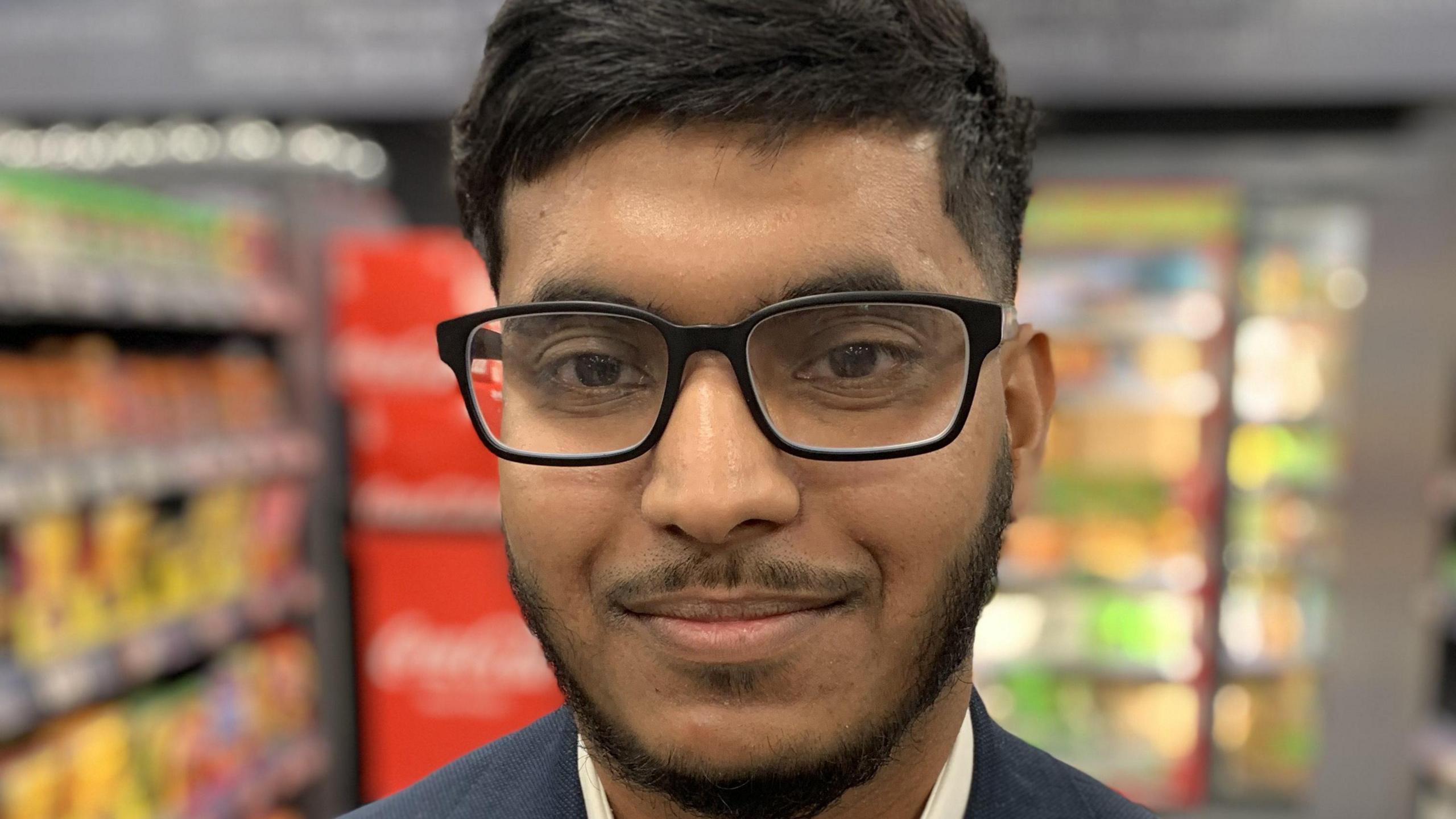 A man stands in a supermarket. He has black hair, a neat black beard and glasses. He is wearing a navy blue jacket and a white shirt. The supermarket shelves in the background are colourful but out of focus.