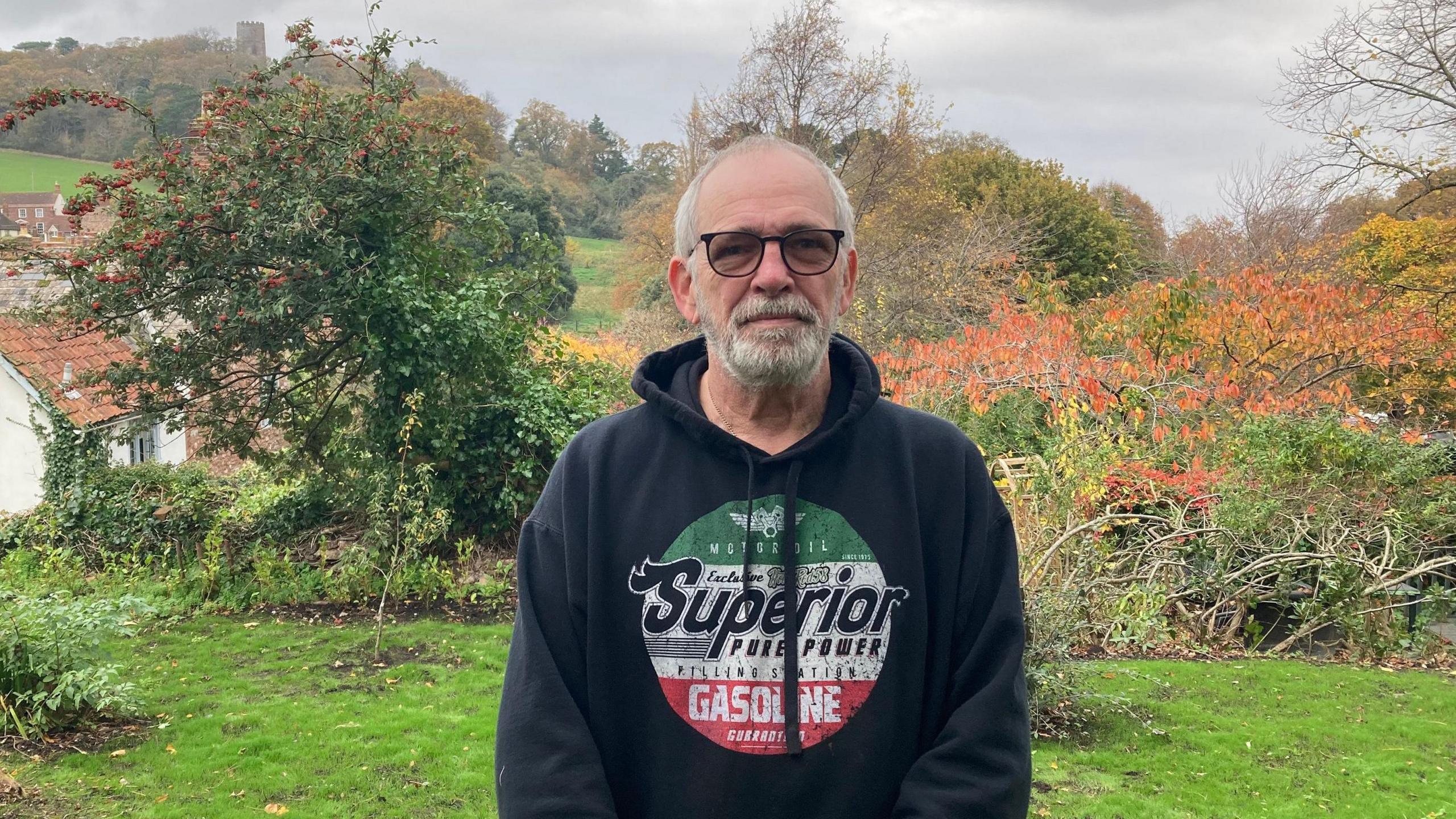 Ian Edmonds wearing a dark hoodie. He has glasses on and a short beard. He is standing on the edge of a small clearing, and you can see Dunster Castle in the background, with a tower just poking over some trees.
