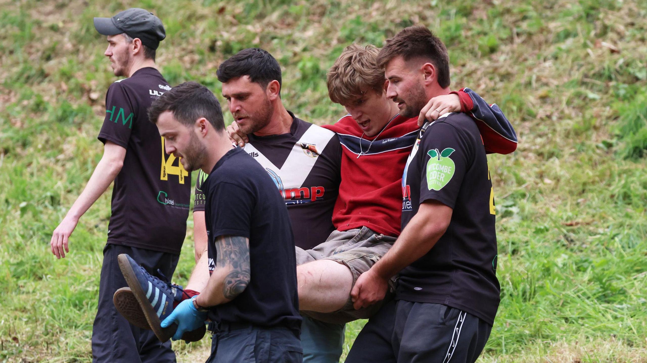 A young man in a red T-shirt and grey shorts is carried away by three male volunteers in black at the end of a race at the Gloucestershire cheese rolling