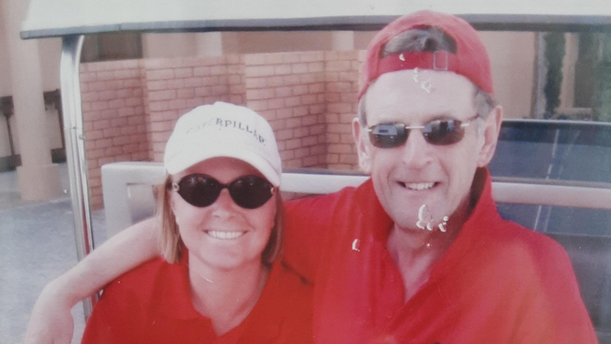 Two people in red t-shirts smiling into the camera with caps on.