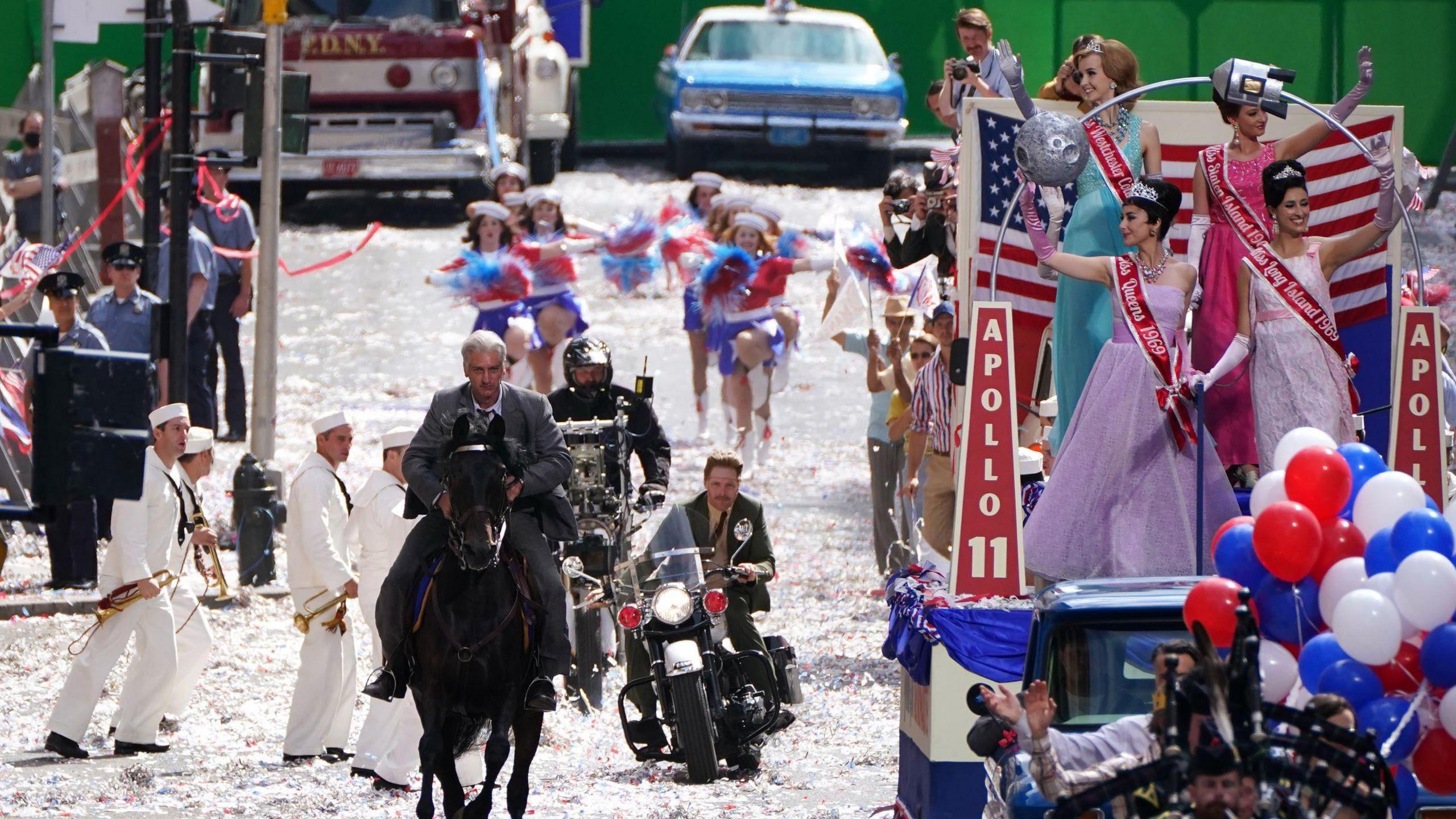 Filming on the last Indian Jones film 0- a stuntman rides a horse while being chased by another man on a motorbike. Sailors are watching them, while a parade continues around them - two women dressed as beauty queens are on one of the floats. 