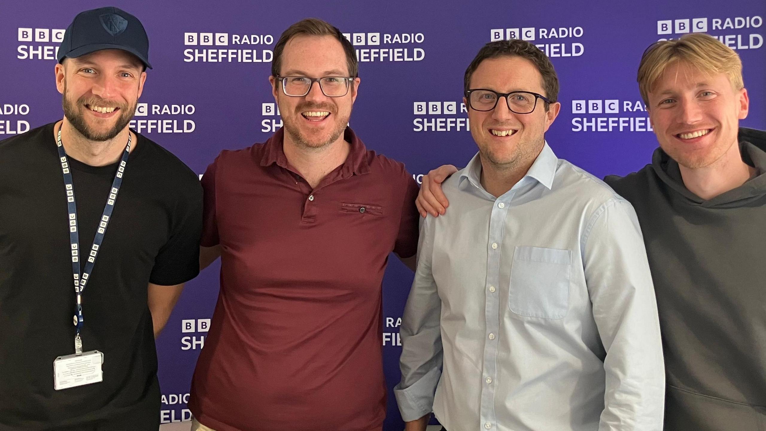 Four men stood in front of a purple BBC Radio Sheffield banner. They are all over 6ft in height and smiling in the pitcure. Two of them have dark hair and glasses, another wears a baseball cap while the fourth man has medium-length blonde hair.