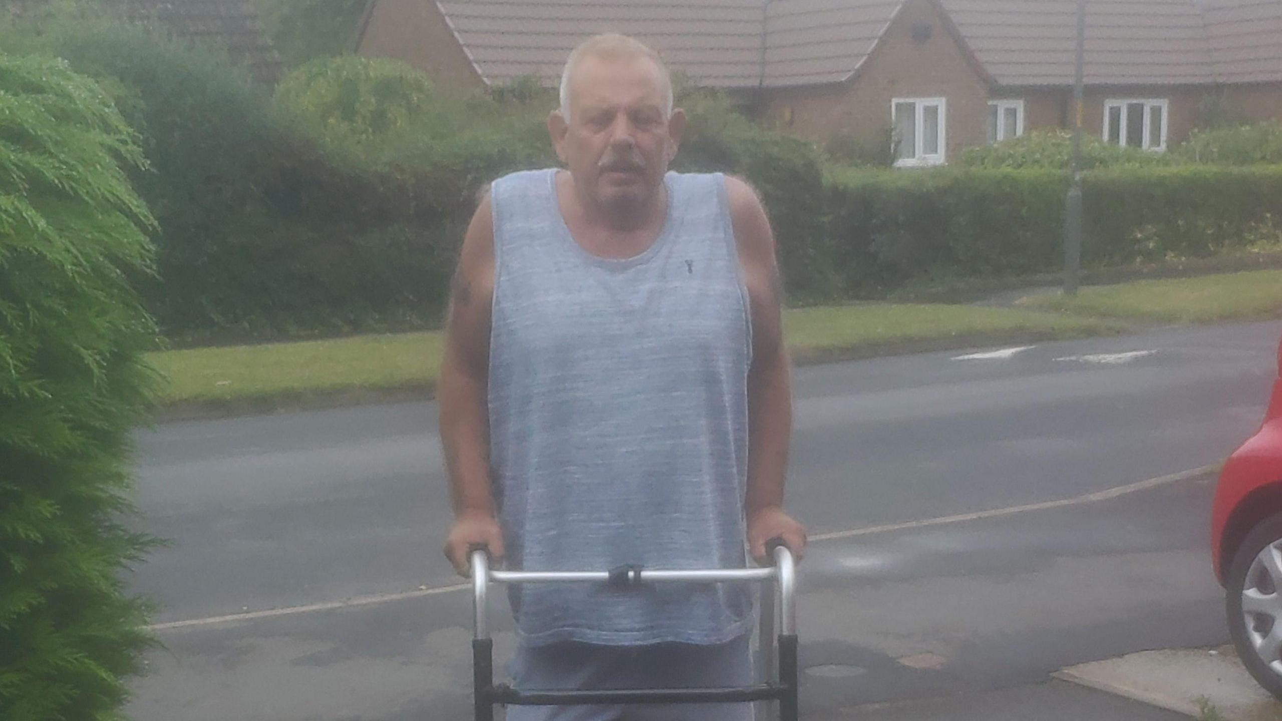 Denny in a grey vest using a walking frame