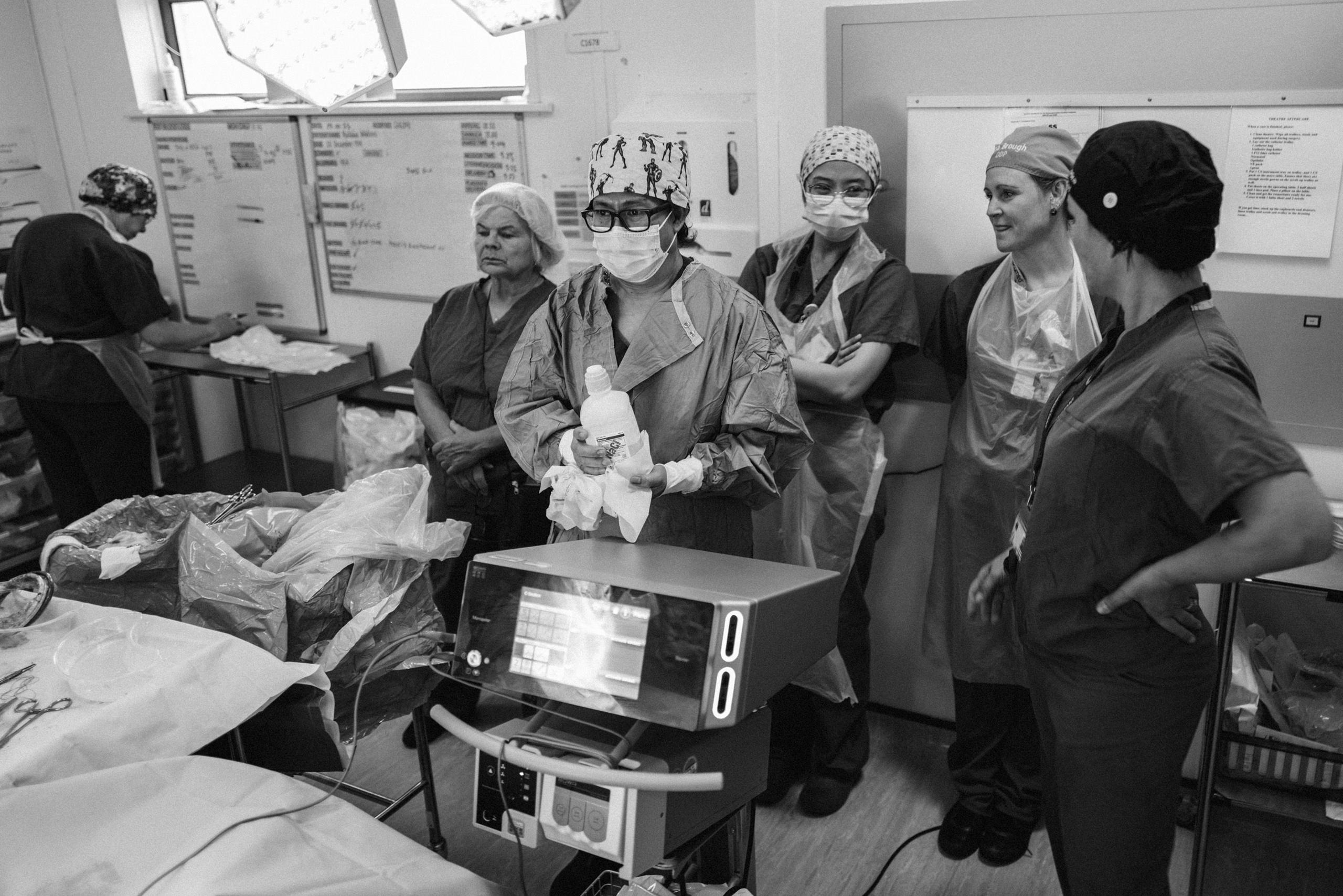 Six staff members are seen standing near the wall of an operating theatre with medical equipment in front of them. All are wearing scrubs with some wearing surgical gowns and masks. A separate staff member is seen working on a table in the corner of the room.