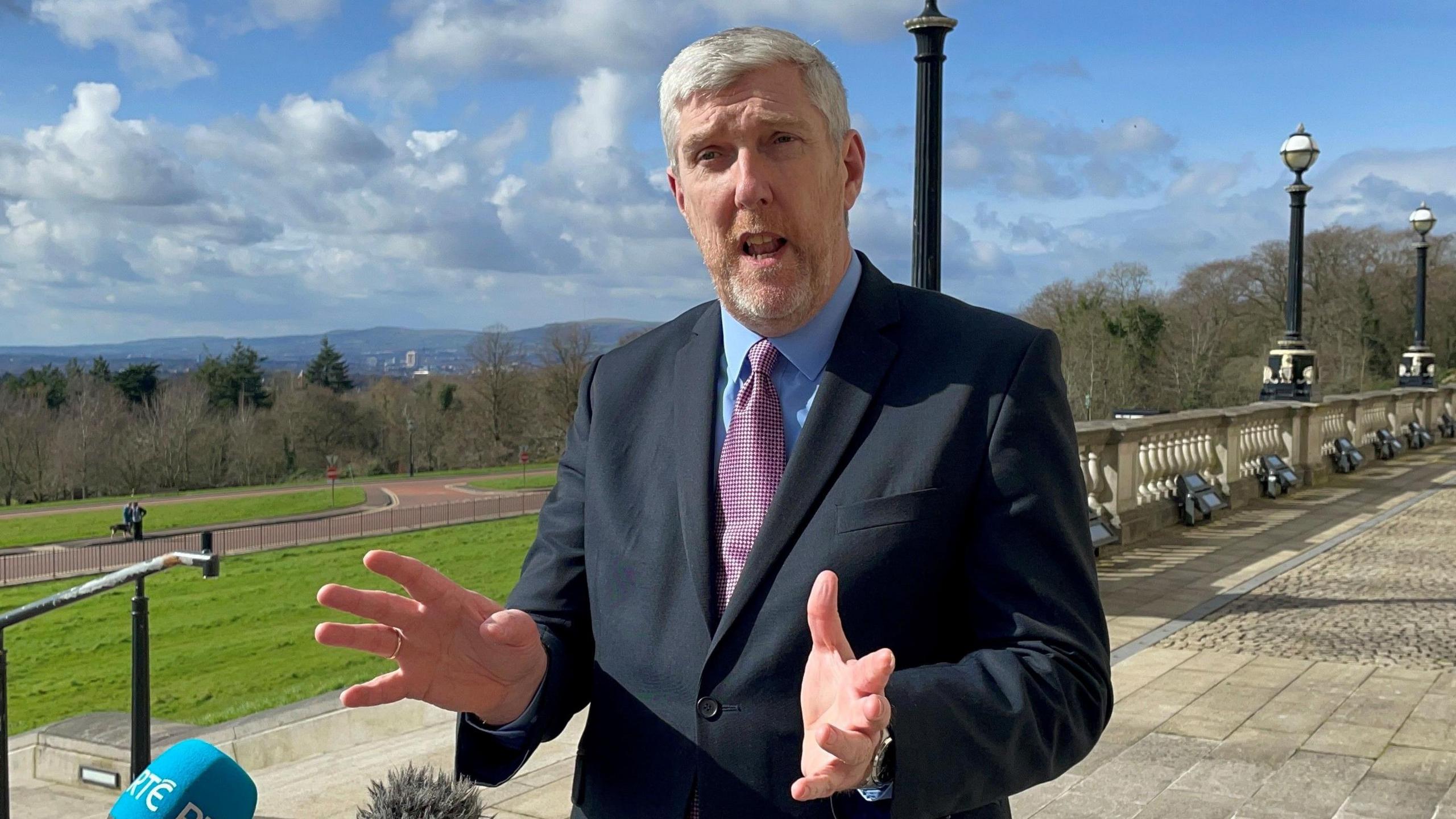 Infrastructure Minister John O'Dowd at Stormont in a navy suit, blue shirt and pink tie, speaking at microphone