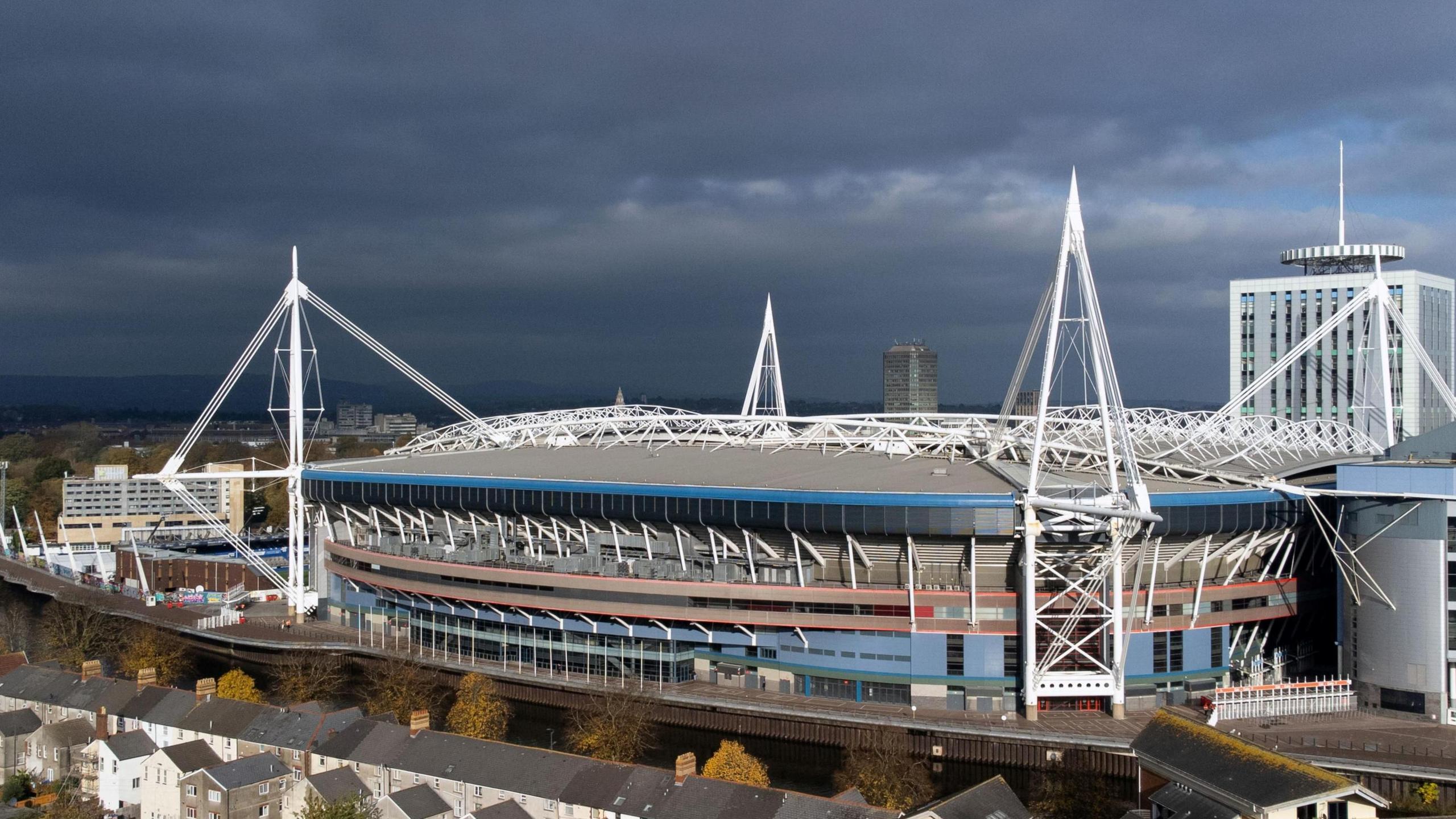 An aerial view of the Principality Stadium. Stormy grey skies are seen behind it, while in front runs the river and trees and a row of terraced houses.