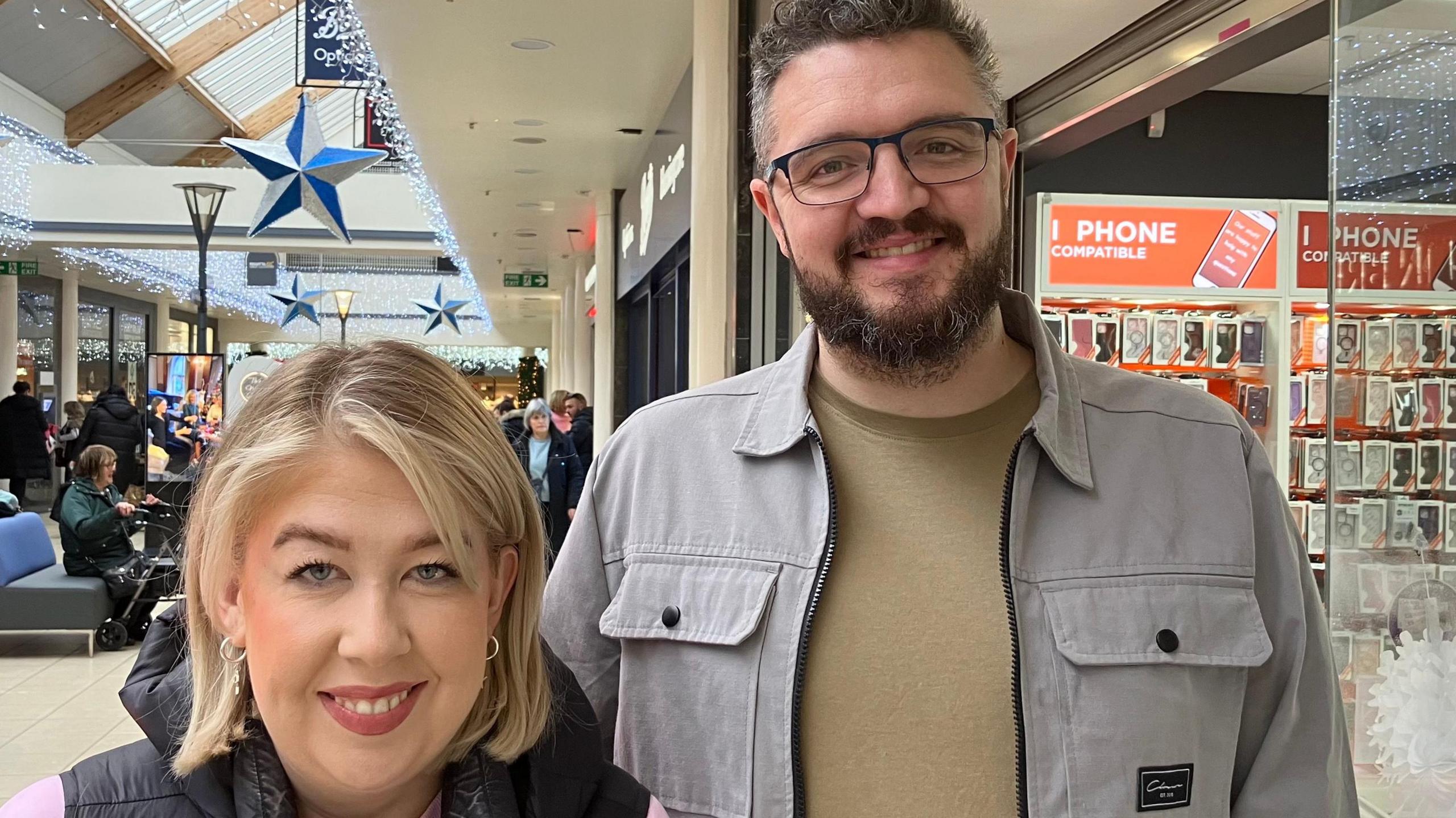 A woman and man stand smiling into the camera.  The blonde haired woman is wearing a pink top under a padded body warmer.  The man beside her who is wearing dark rimmed glasses has a short beard.  He is wearing a fawn coloured top under a grey denim looking jacket.