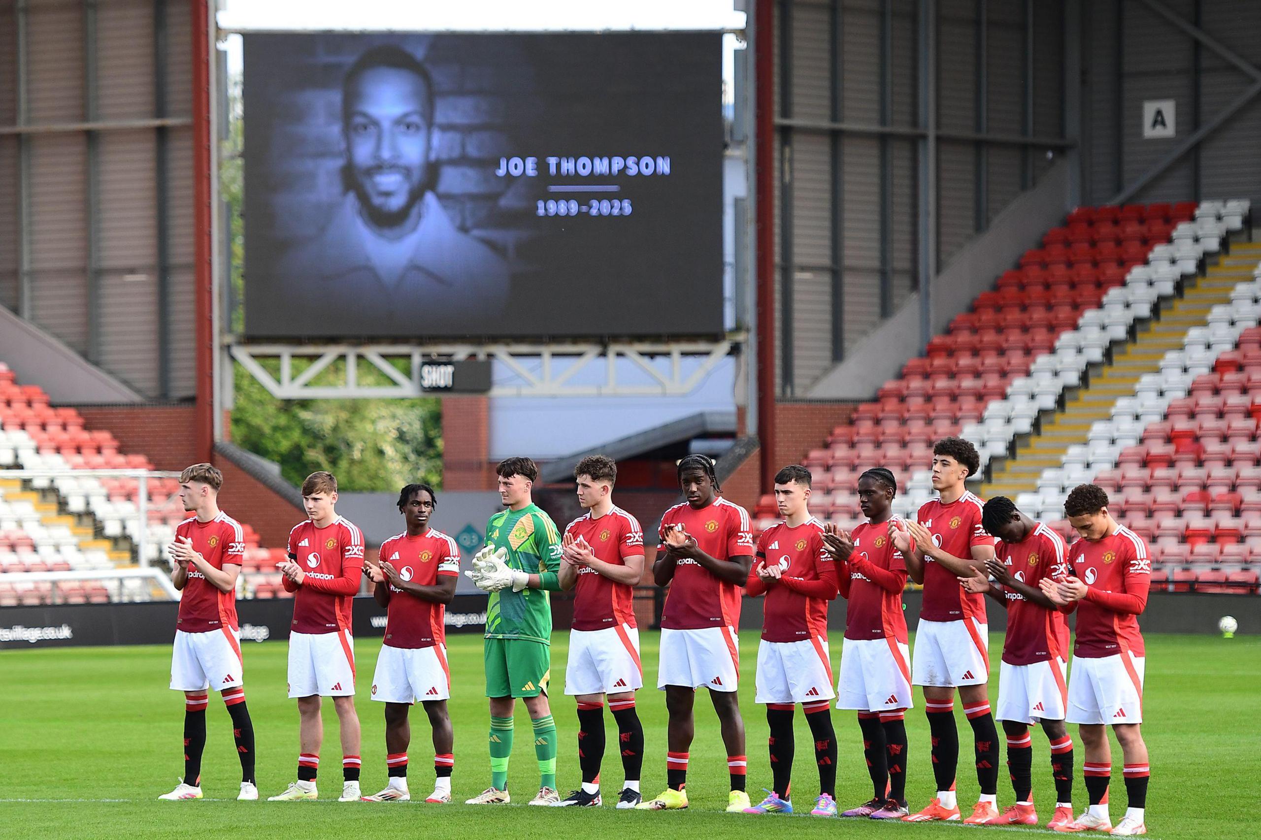 Manchester United academy players doing a minute's applause in memory of Joe Thompson before a game in April. There is a picture of Thompson on a screen behind them.