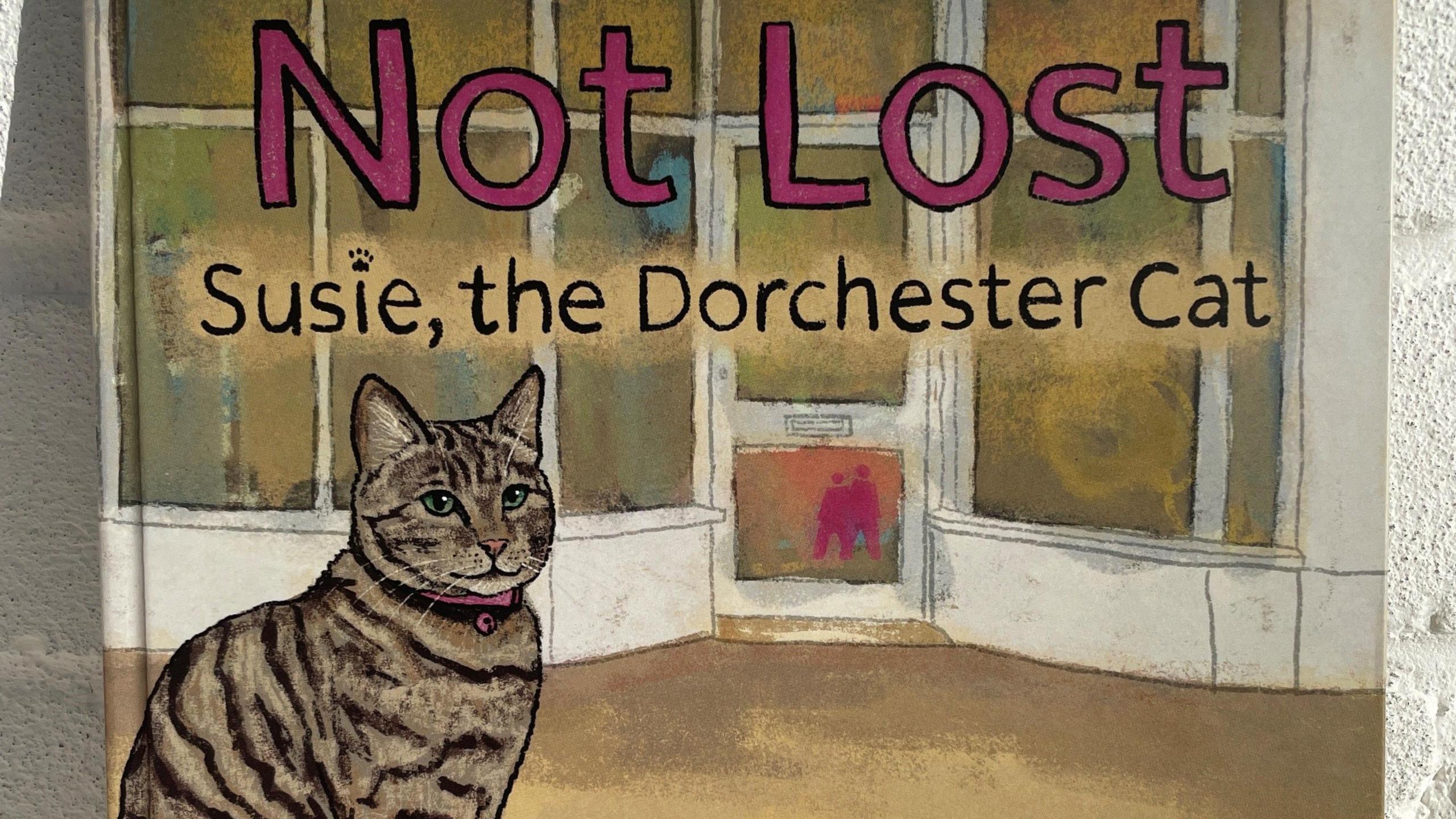 Detail of the book cover with an illustration of the tabby cat sitting in front of a white shop entrance with bow windows. The title reads: Not Lost, Susie the Dorchester Cat.