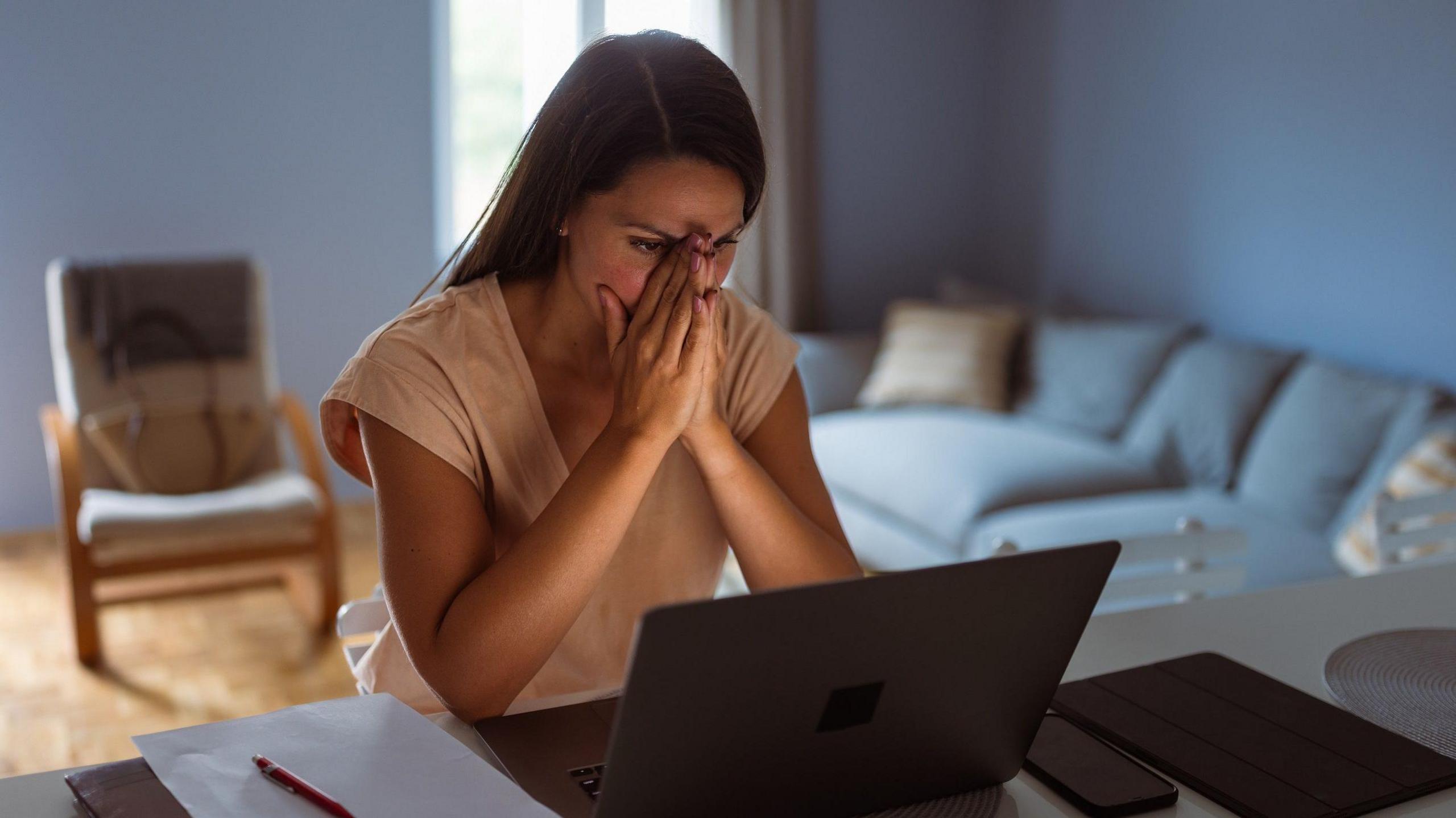 A woman sits at a desk looking at her laptop. She has her head in her hands