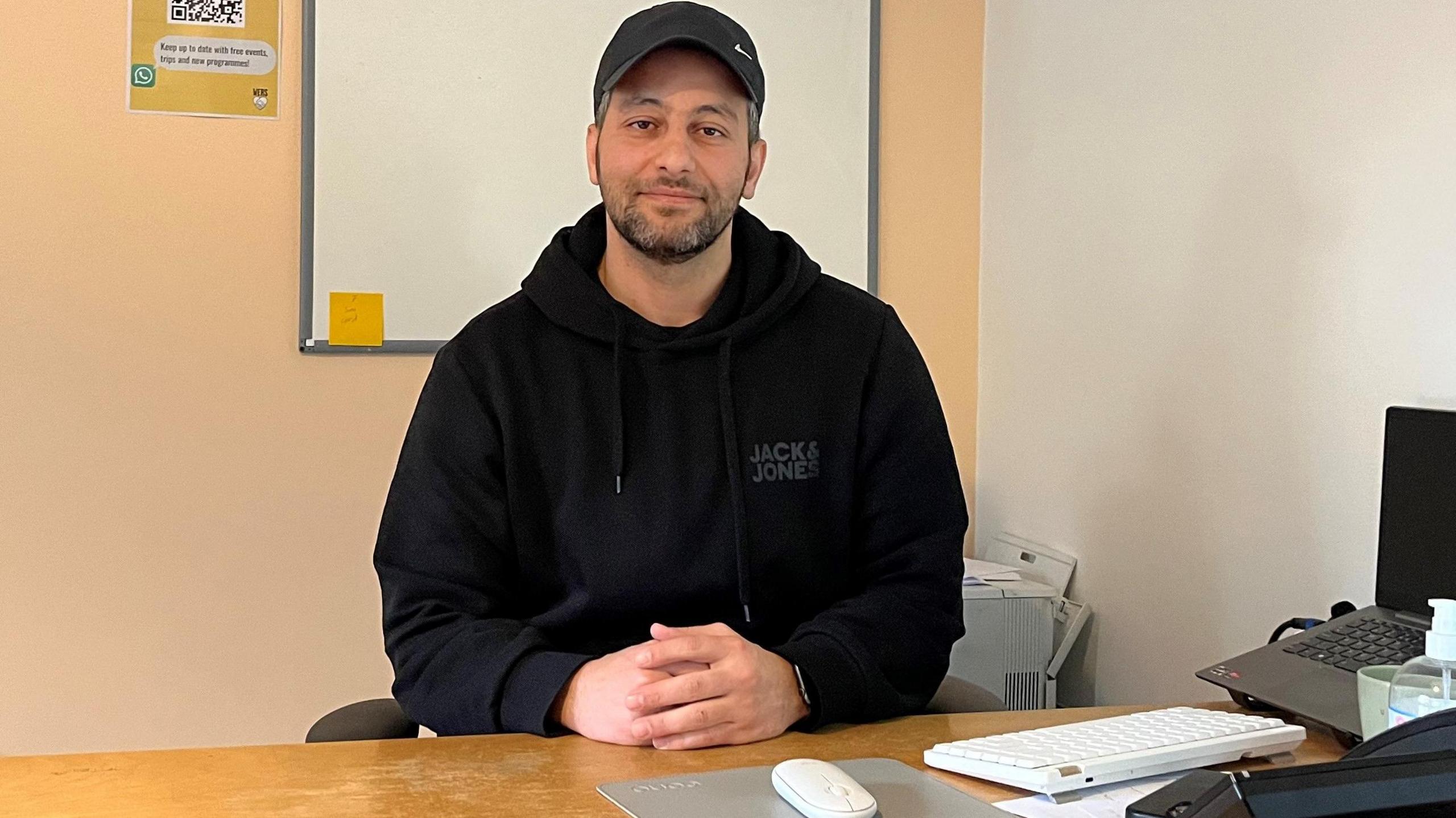 Tarik Alali in a baseball cap and black hoodie sitting by desk with hands clasped. He is looking at the camera. 