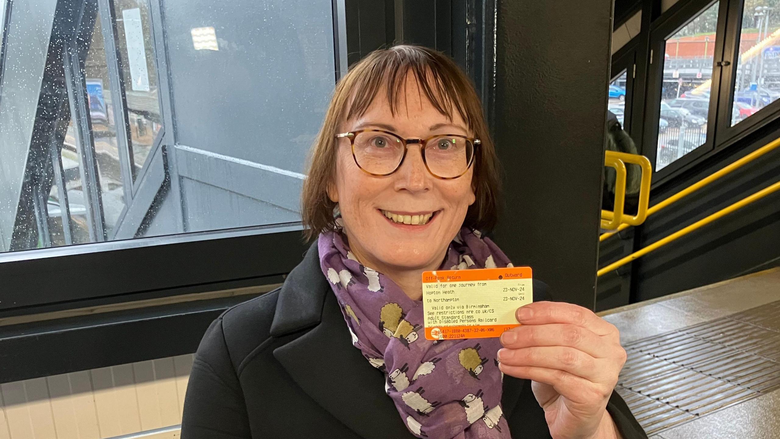 Sarah Ward holding up a train ticket in a railway station. She has short brown hair, is wearing glasses and has a lilac scarf 