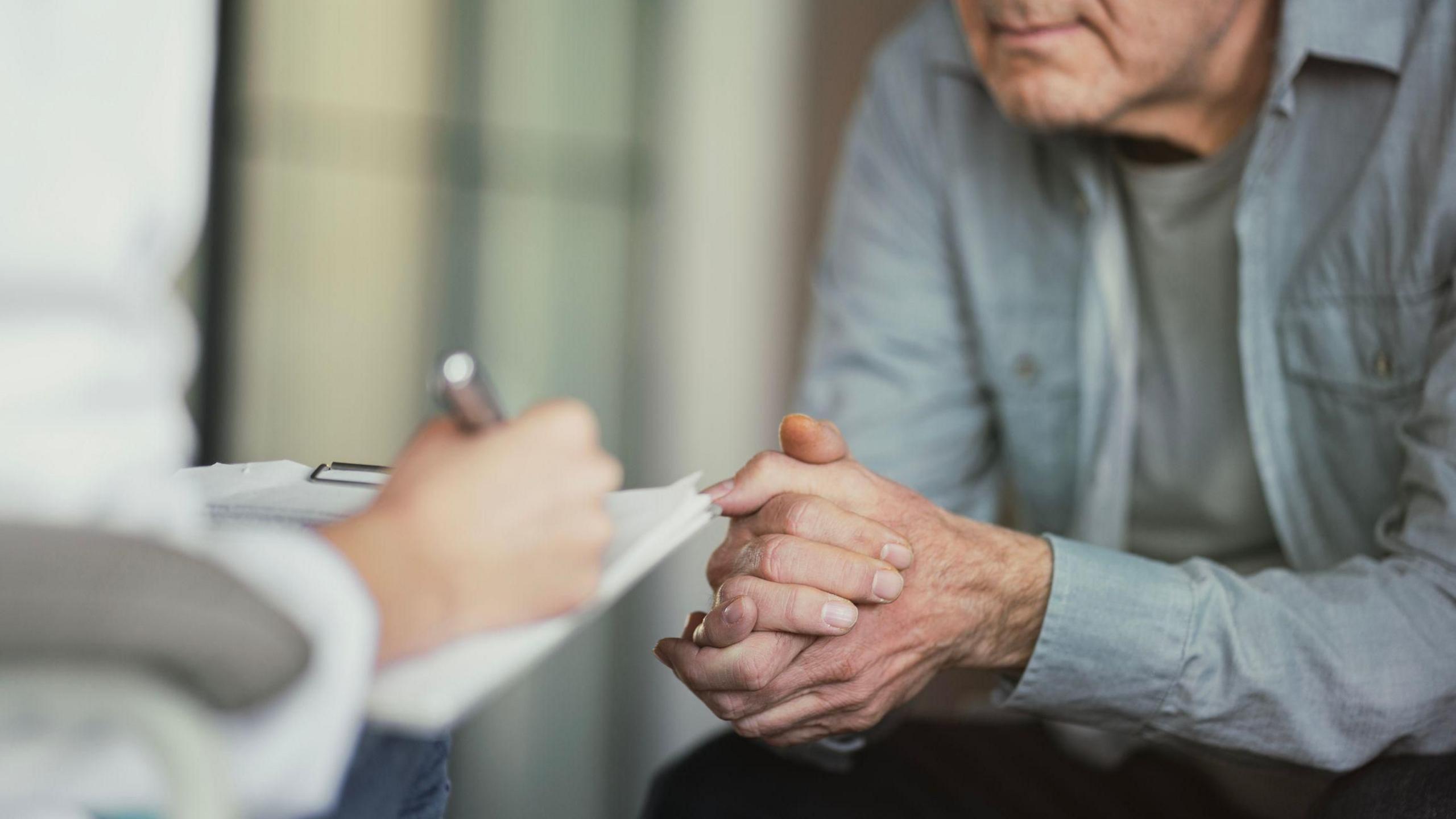 A doctor writes on a clipboard as a patient, sitting with hands clasped, listens