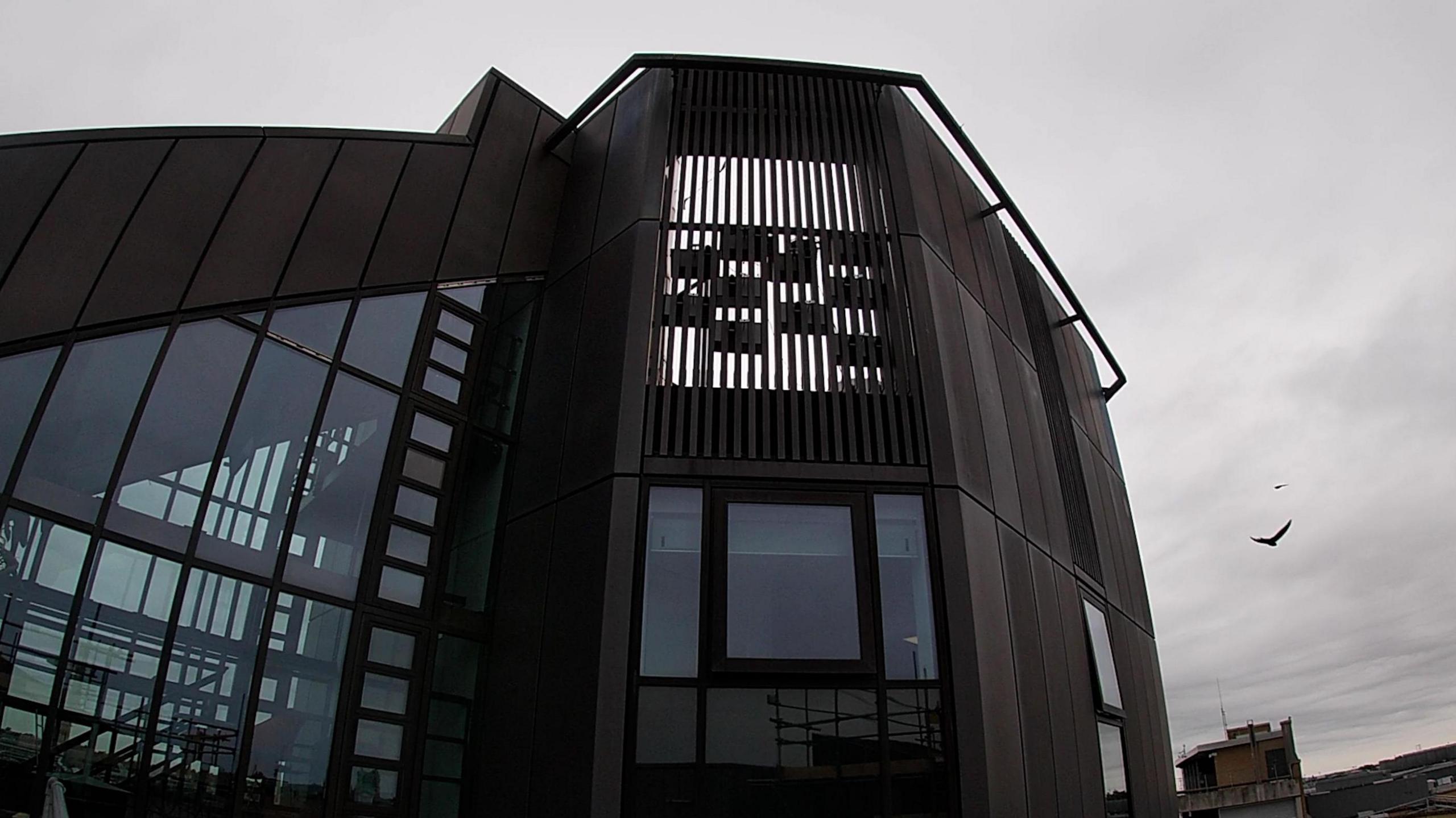 Swift boxes are placed high up on a modern building silhouetted against a grey sky. Birds can be seen flying above.