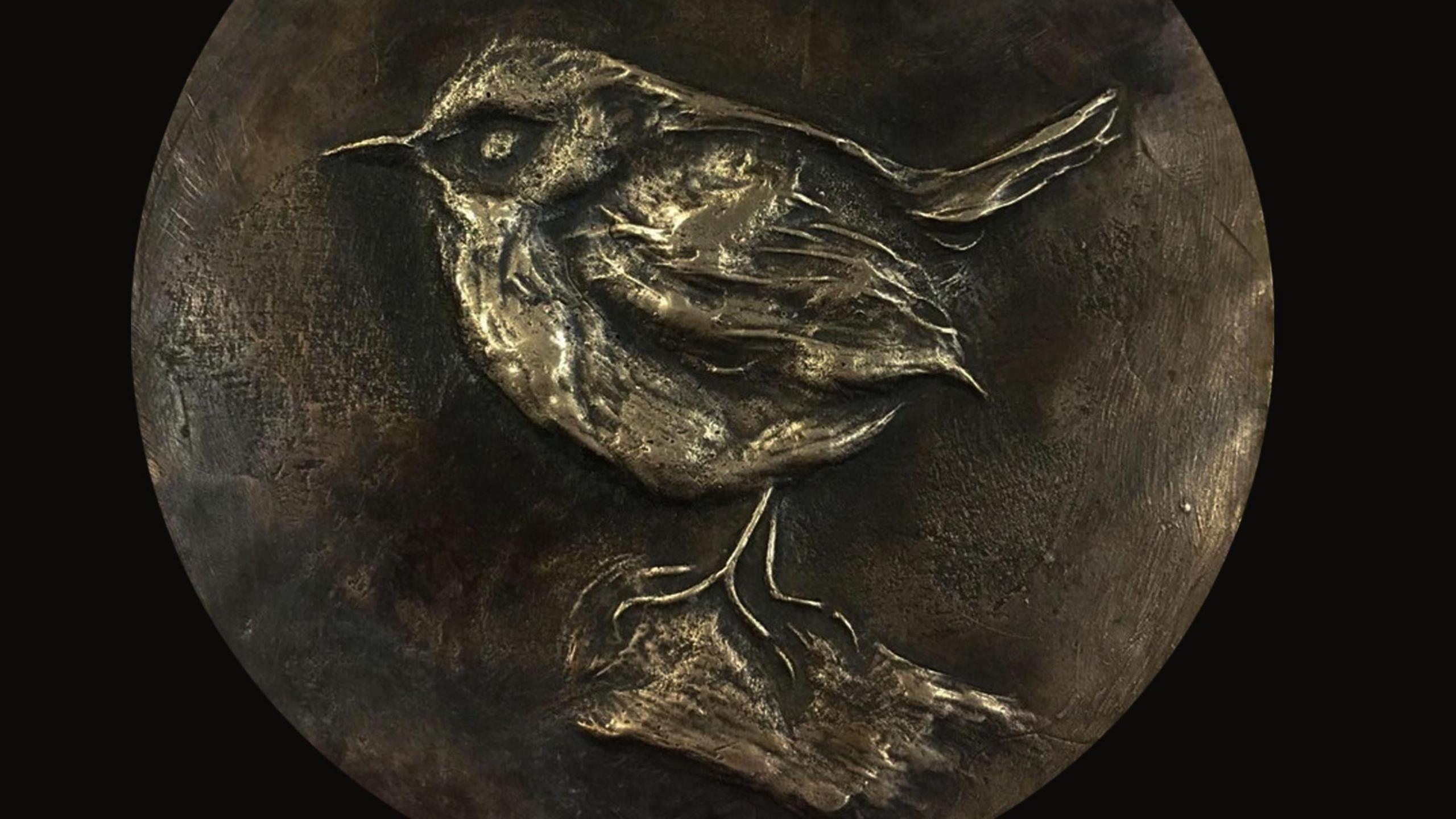 A round piece of gold-coloured metal with a bird engraved in it. The backgrounder is much darker.