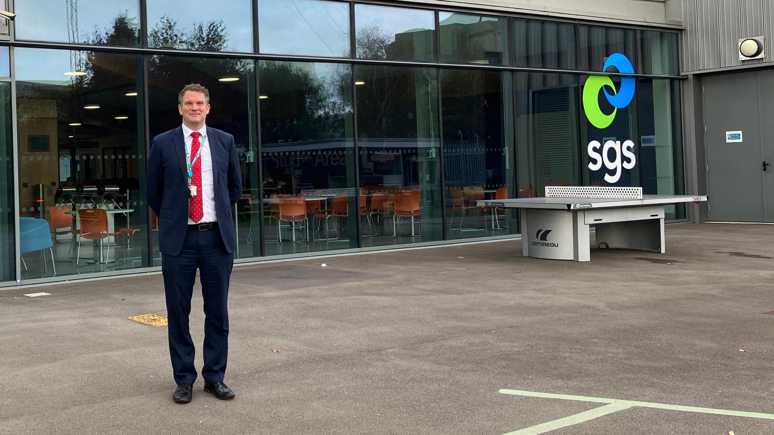 Gareth Lister stood outside of SGS Berkeley Green UTC. The college's blue and green curved logo is visible on the building exterior. Mr Lister is wearing a dark blue suit with a white shirt and bright red tie