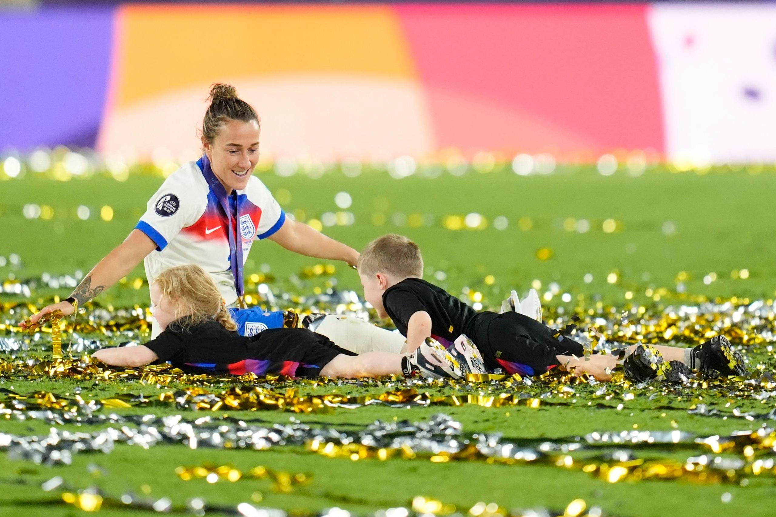 lucy bronze smiles while sat on the stadium pitch which is covered in metallic confetti while two children in tournament kit lie next to her celebrating