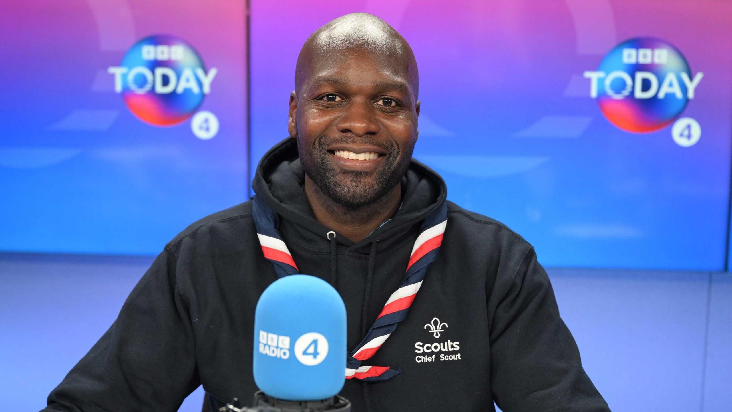 Chief scout Dwayne Fields in the Today studio. A light blue Radio 4 microphone can be seen in the foreground in front of him and the Today logo can be seen on screens in the background. Fields is wearing his scout necker on top of his black hoodie which has the Scouts logo and his title printed on the left breast.