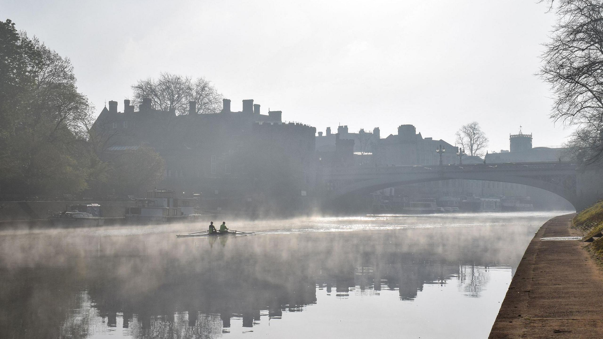 The River Ouse taken in the morning, with mist over the water and it is just getting light. Two people are rowing in the middle of the water and just their silhouettes are visible.