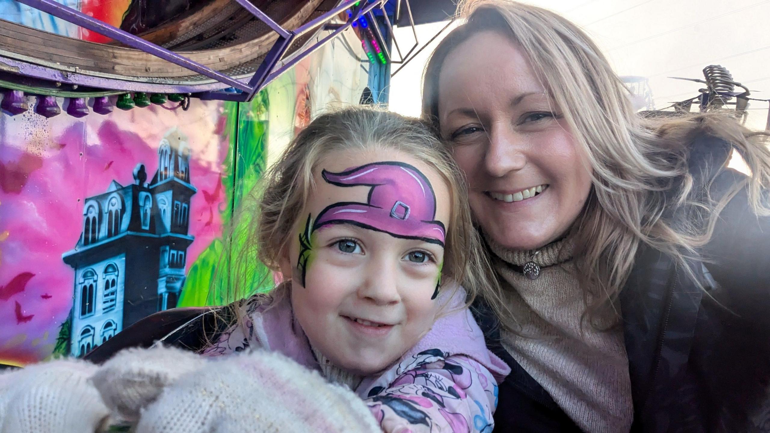 Tracey Watkins with her arm around her daughter aged about 5. The girl has face paint on her forehead and temples and a fairground attraction is visible in the background