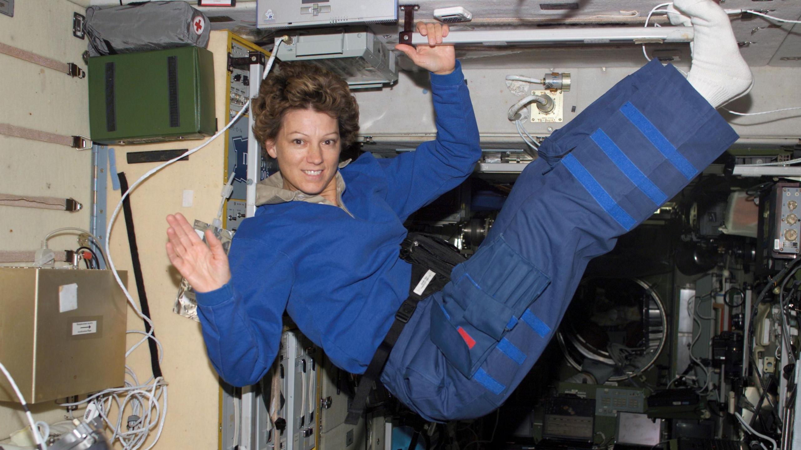 Eileen Collins in mid air in a spacecraft. She is smiling and waving to the camera and her legs are raised in the air. She is wearing a blue top and trousers with white socks. She is holding on to a bar with her left hand and is surrounded by wires and electronic hardware.