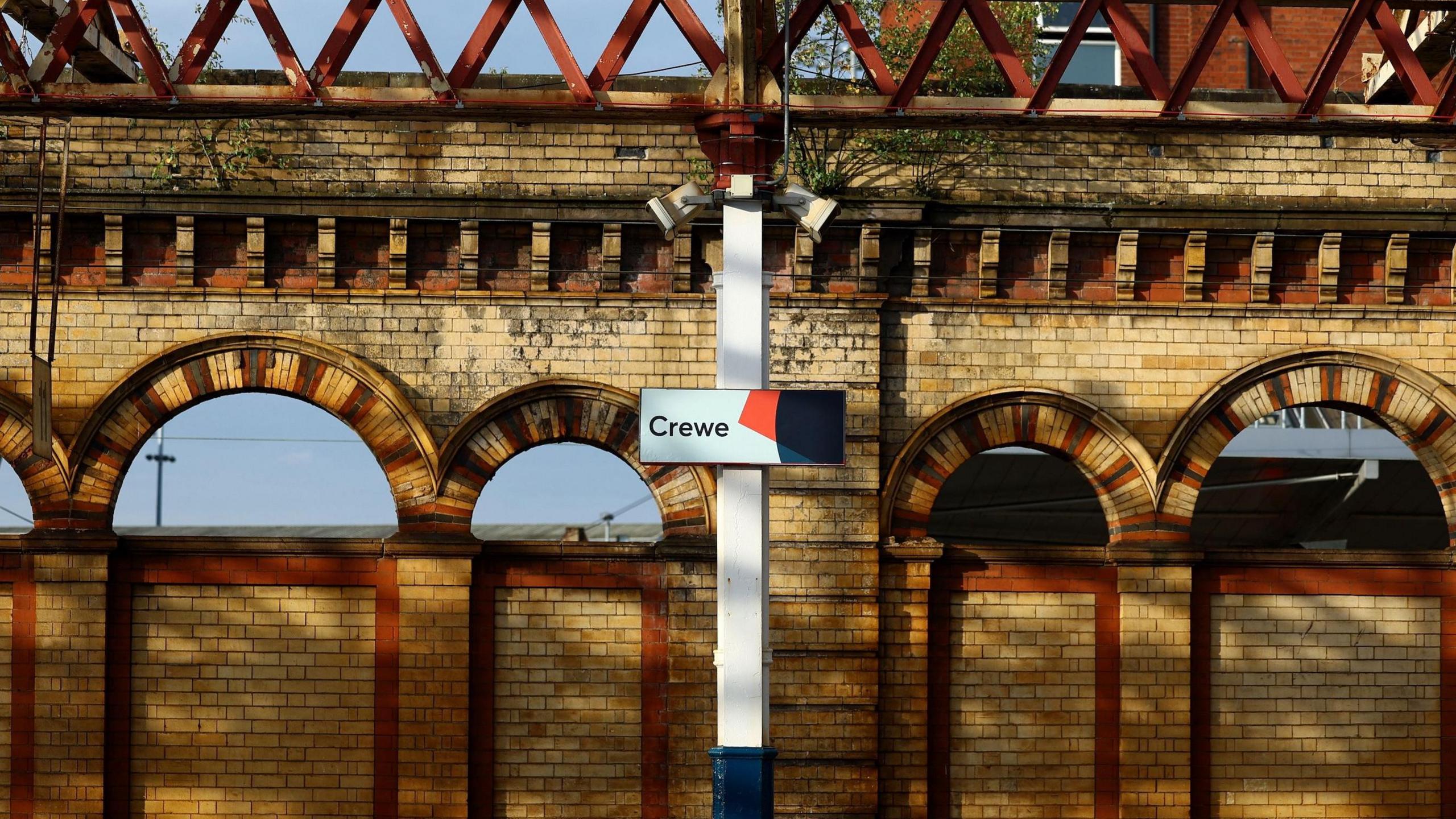 A view of a sign saying Crewe inside the railway station. The sign sits in front of a row of arches in light brick with orange and black insets.