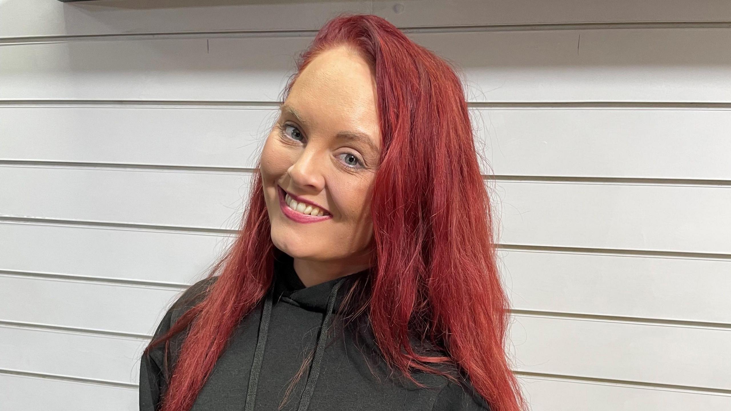 Jennie Doolan is smiling directly at the camera. She is wearing a black hooded jumper and is standing in front of a white panelled wall.